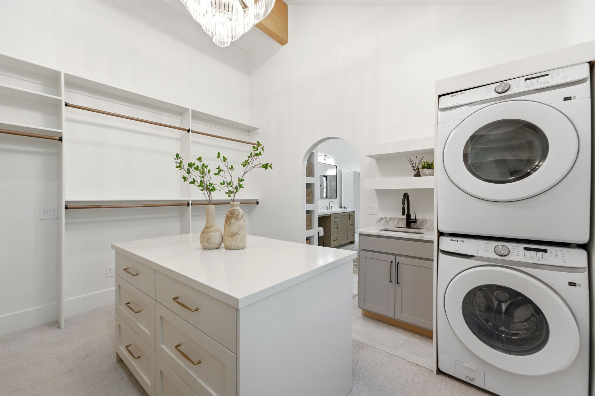 Spacious walk-in closet featuring custom built-in shelving and hanging rods, a central island with drawers, a stacked washer and dryer, and a utility sink with a matte black faucet