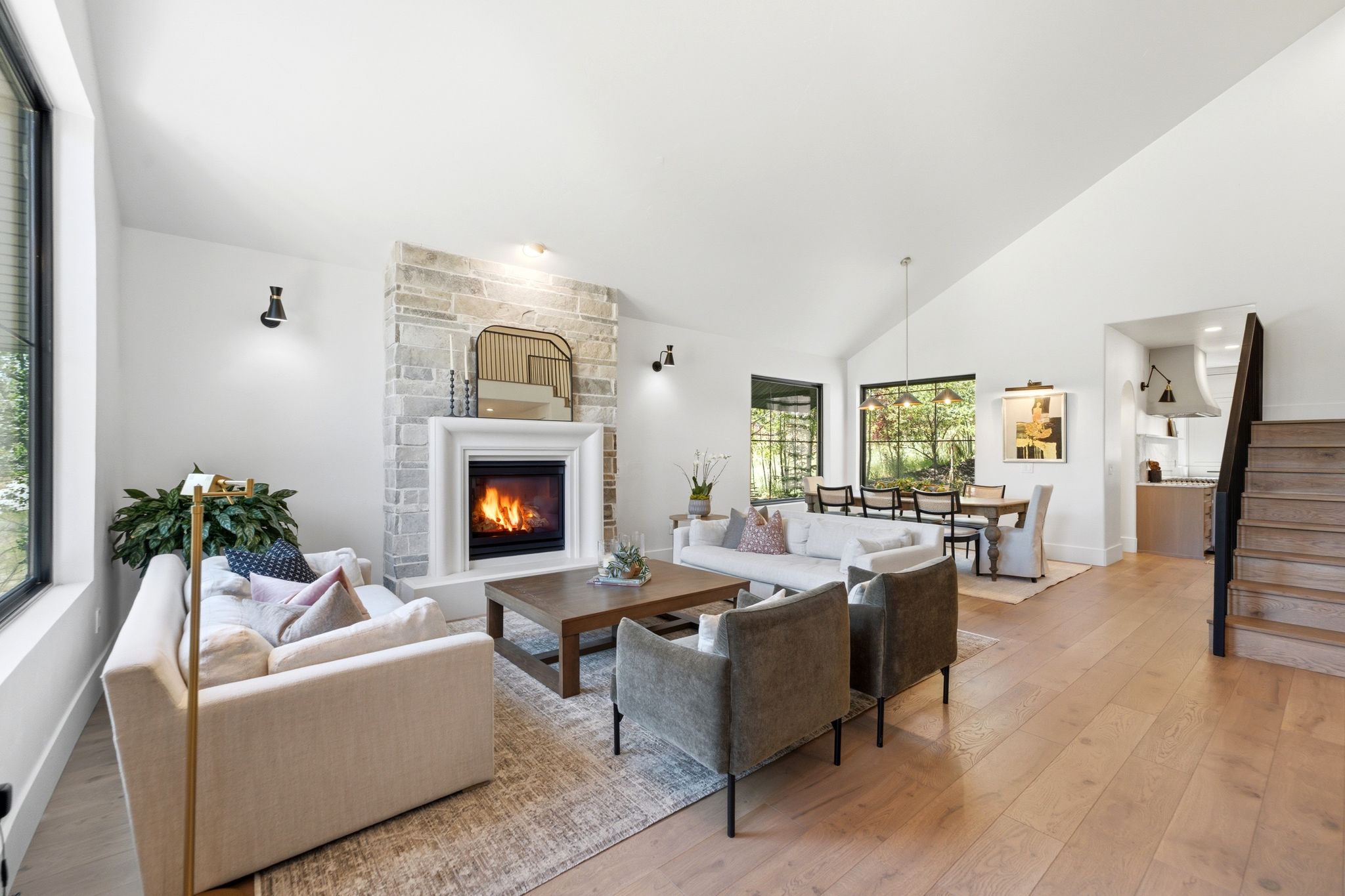 Expansive living area featuring a stone-clad fireplace with a white mantel, wide plank wood-finish flooring, soaring vaulted ceilings, and large windows