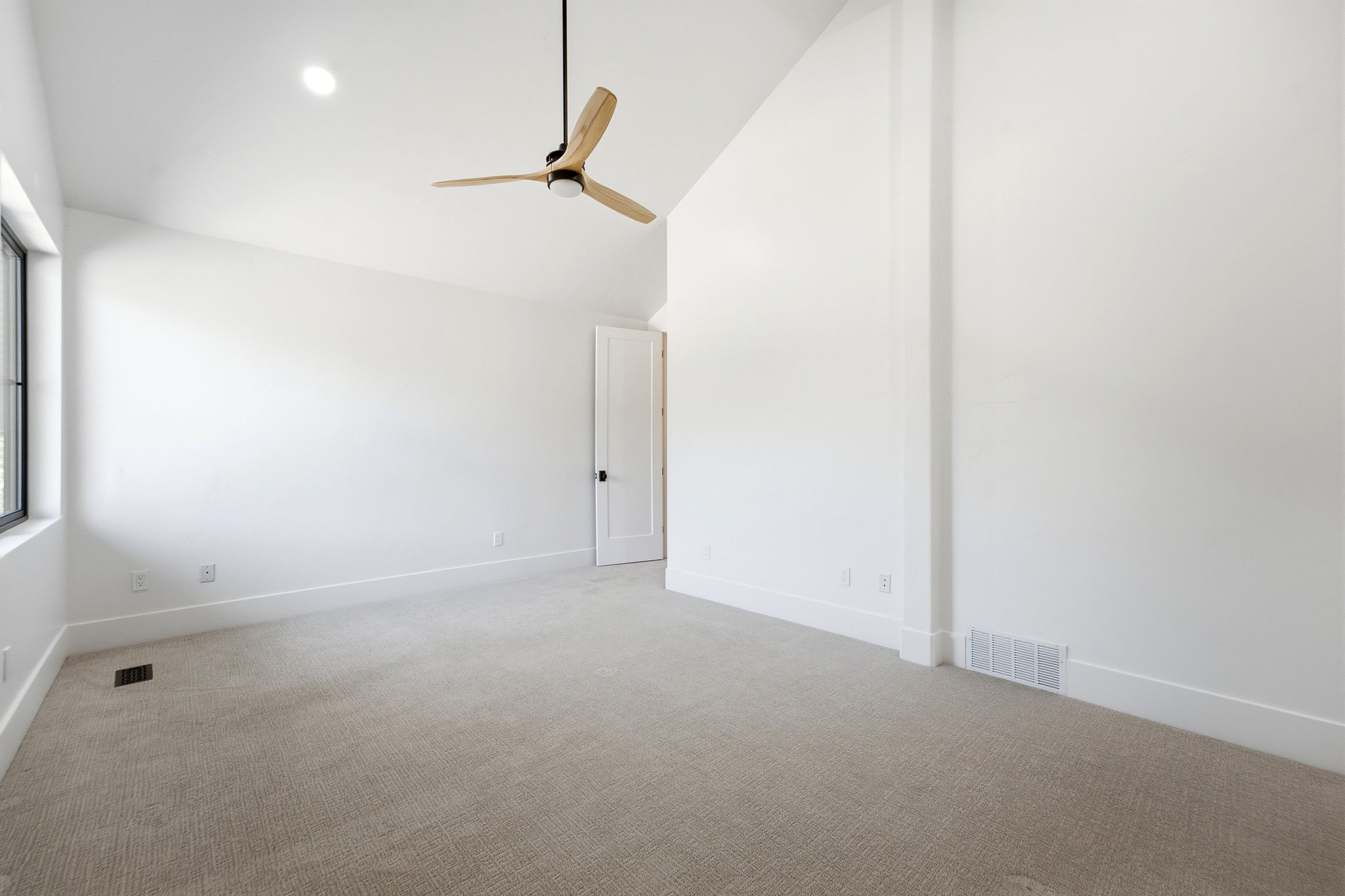 Spacious interior with vaulted ceilings, featuring neutral carpeting, white trim, a modern ceiling fan, and a single window with black framing