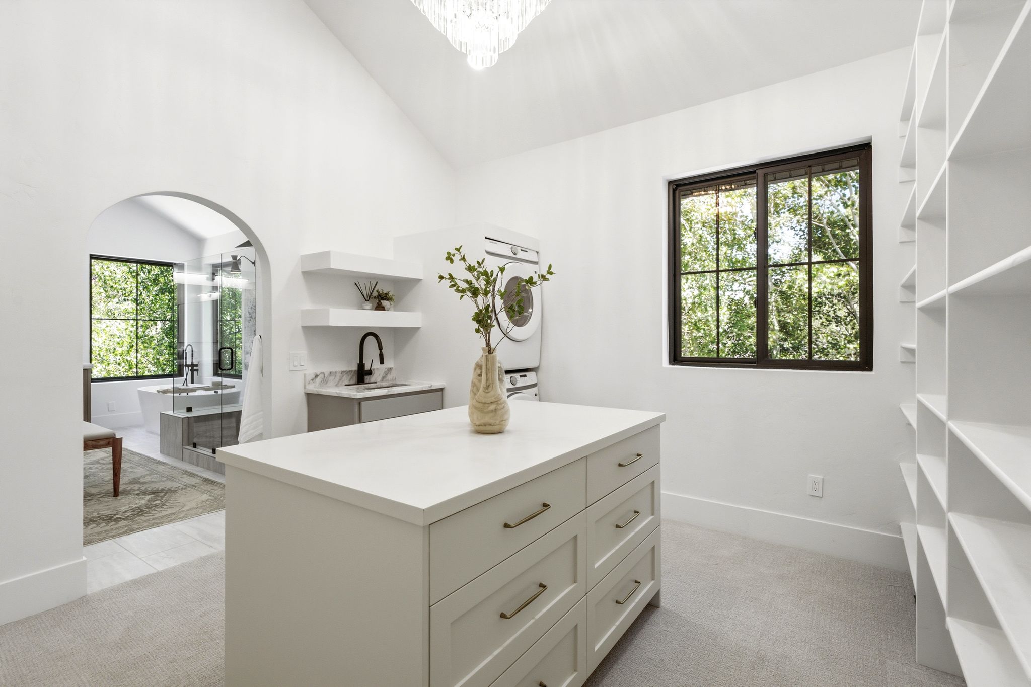 Spacious dressing room featuring a central island with drawers, extensive built-in shelving, a laundry area with stacked appliances, and an arched entry to a bathroom with a freestanding tub