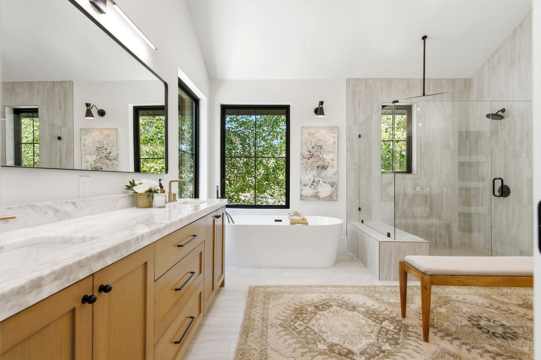 Spacious bathroom featuring a double vanity with light wood cabinetry and a marble-look countertop, an oval freestanding soaking tub, a frameless glass shower with a built-in bench, and multiple windows with black trim