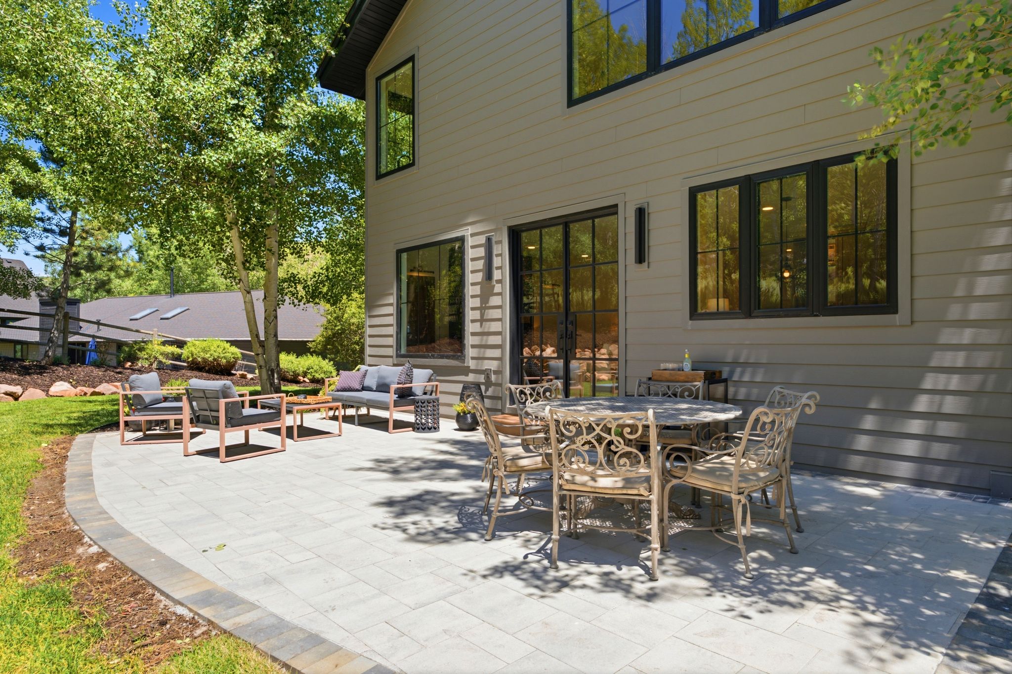 Expansive outdoor patio featuring paver stone hardscaping, multiple seating areas, a glass-paned exterior door, and black-framed windows