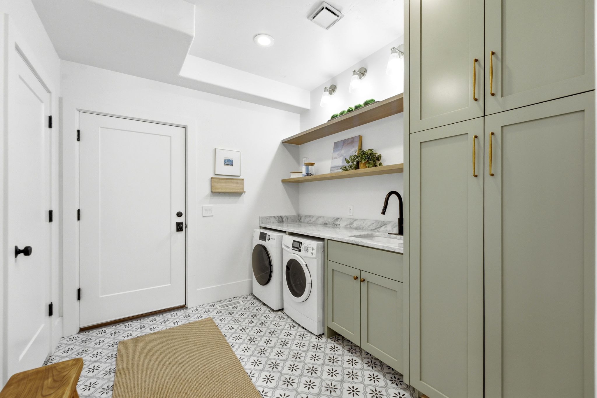 Laundry room featuring patterned floor tiles, light green cabinetry with gold-tone hardware, a sink with a matte black faucet, open wood shelving, and white doors with matte black hardware