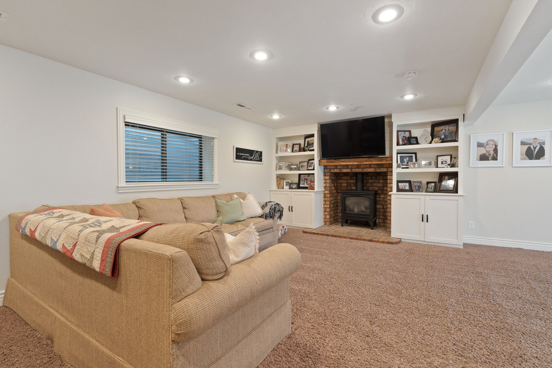 Living area featuring a brick-surround fireplace with a black wood-burning stove, built-in shelving, white cabinetry, recessed lighting, and a light-colored window with blinds
