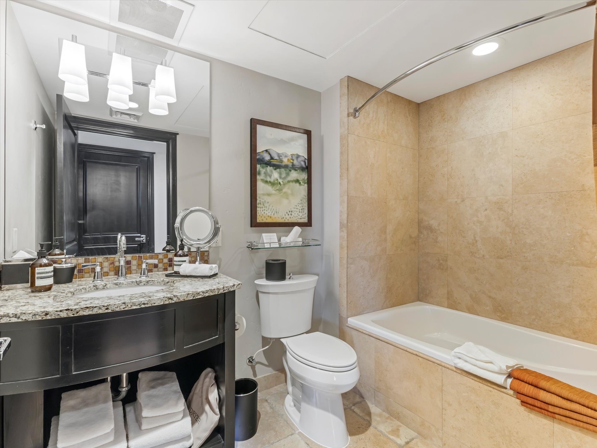Bathroom featuring a built-in bathtub with floor-to-ceiling tiled surround, a dark wood-finish vanity with granite countertop and undermount sink, a mosaic tile backsplash, and a wall-mounted mirror with overhead lighting