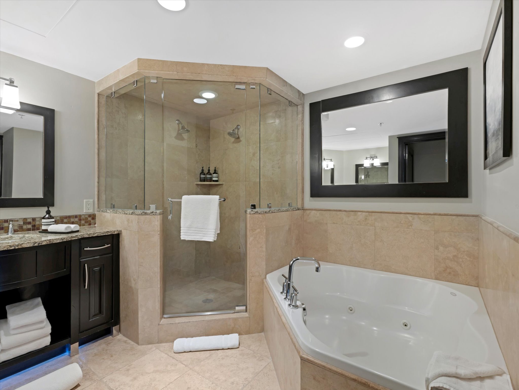 Bathroom featuring a corner jetted soaking tub, a spacious glass-enclosed shower with multiple showerheads, and a vanity with a stone-finish countertop
