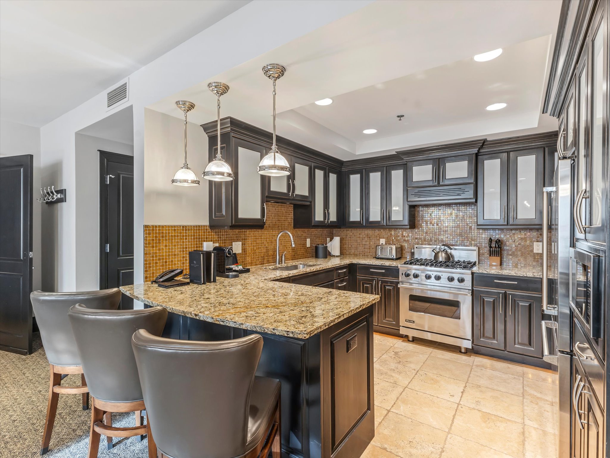Gourmet kitchen featuring dark wood cabinetry, stainless steel appliances, a mosaic tile backsplash, a large breakfast bar with pendant lighting, and neutral-toned tile flooring