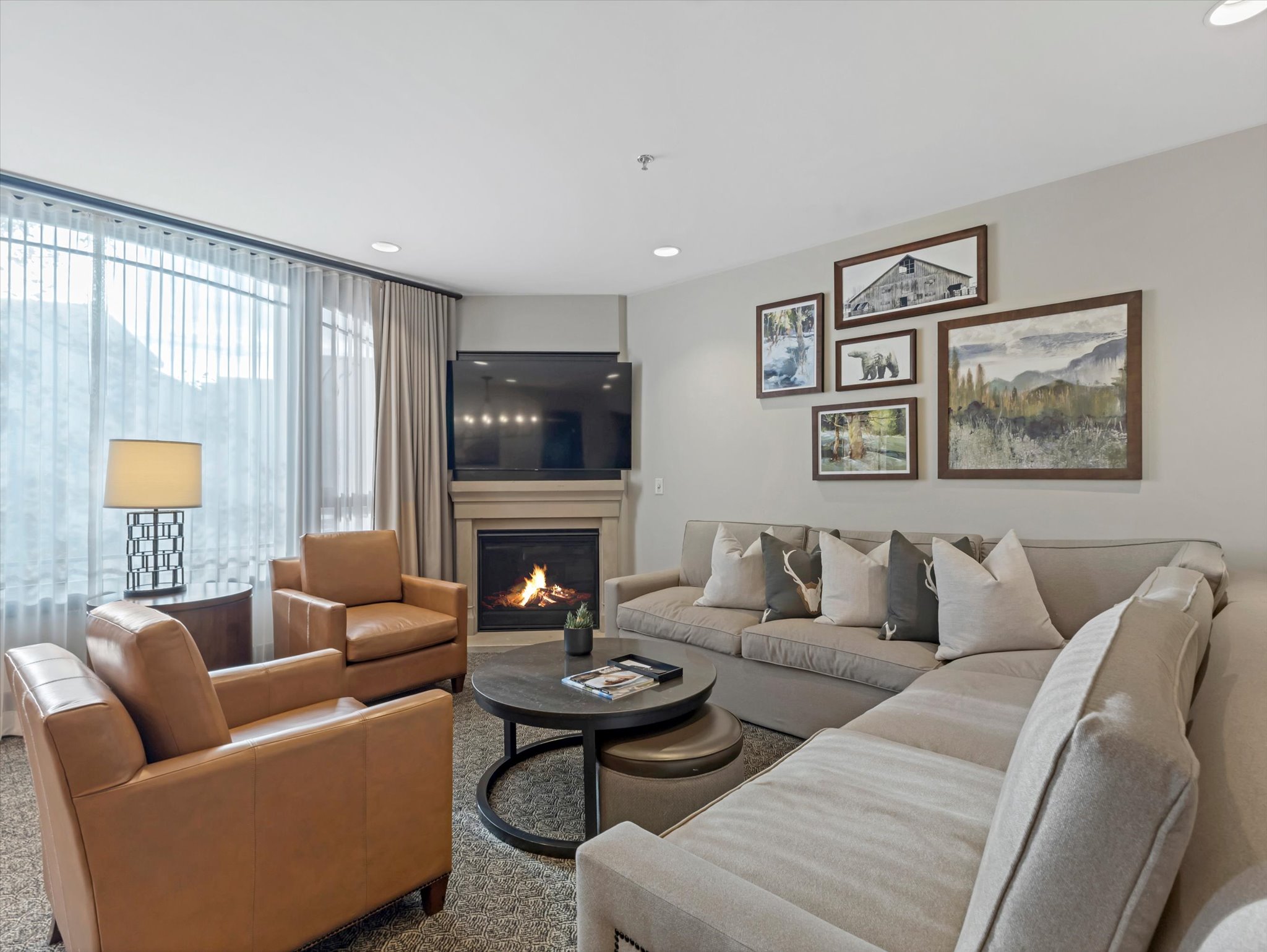Living area featuring a corner fireplace with a stone surround, wall-mounted television, large window with sheer and blackout drapes, recessed lighting, and carpet flooring