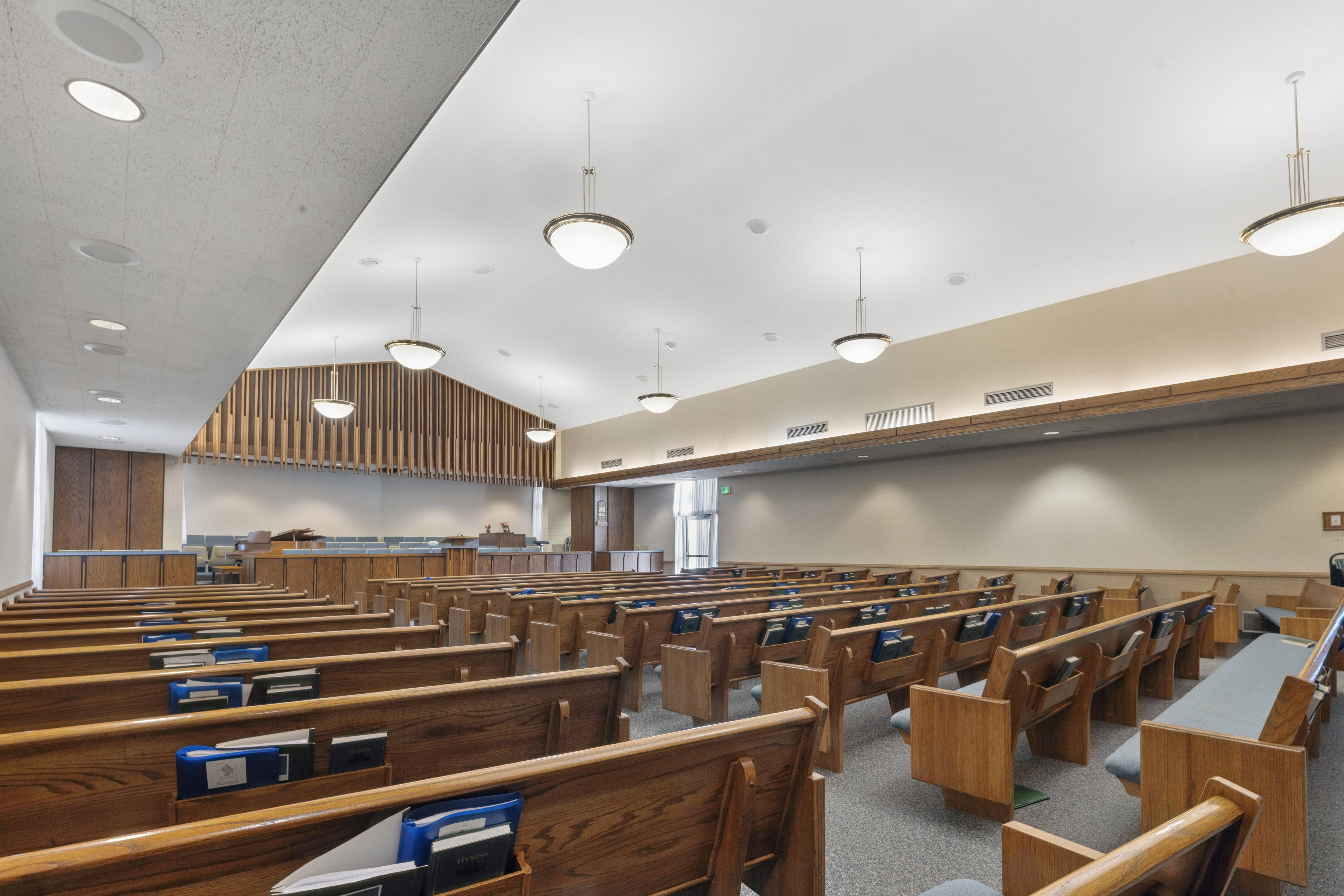 Spacious interior featuring rows of wood benches, acoustic paneling, suspended globe pendant lighting, recessed ceiling fixtures, and neutral-toned walls