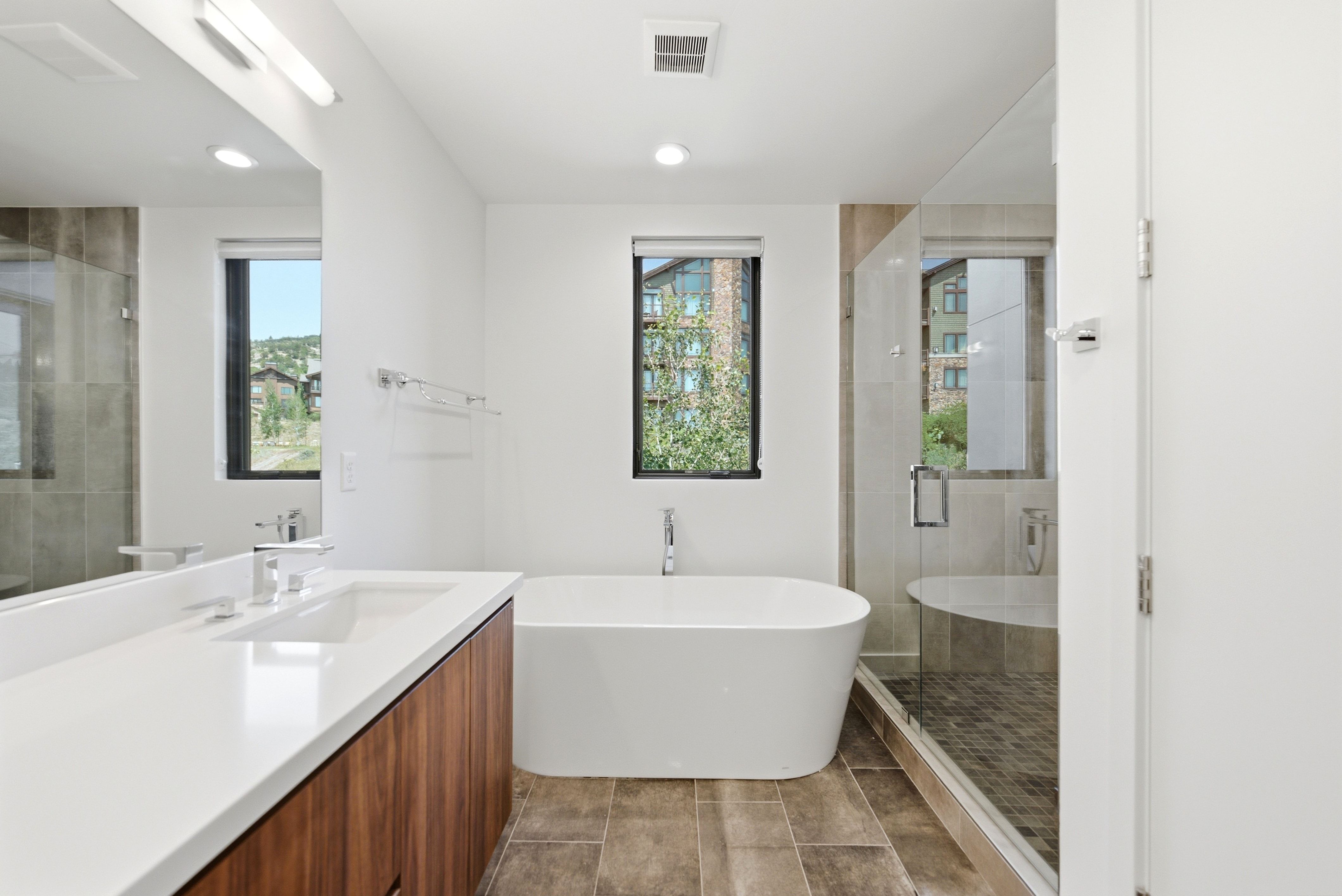 Freestanding soaking tub with a floor-mounted faucet, glass-enclosed shower, dual vanity with a white countertop, wood-finish cabinetry, and contemporary rectangular floor tiles