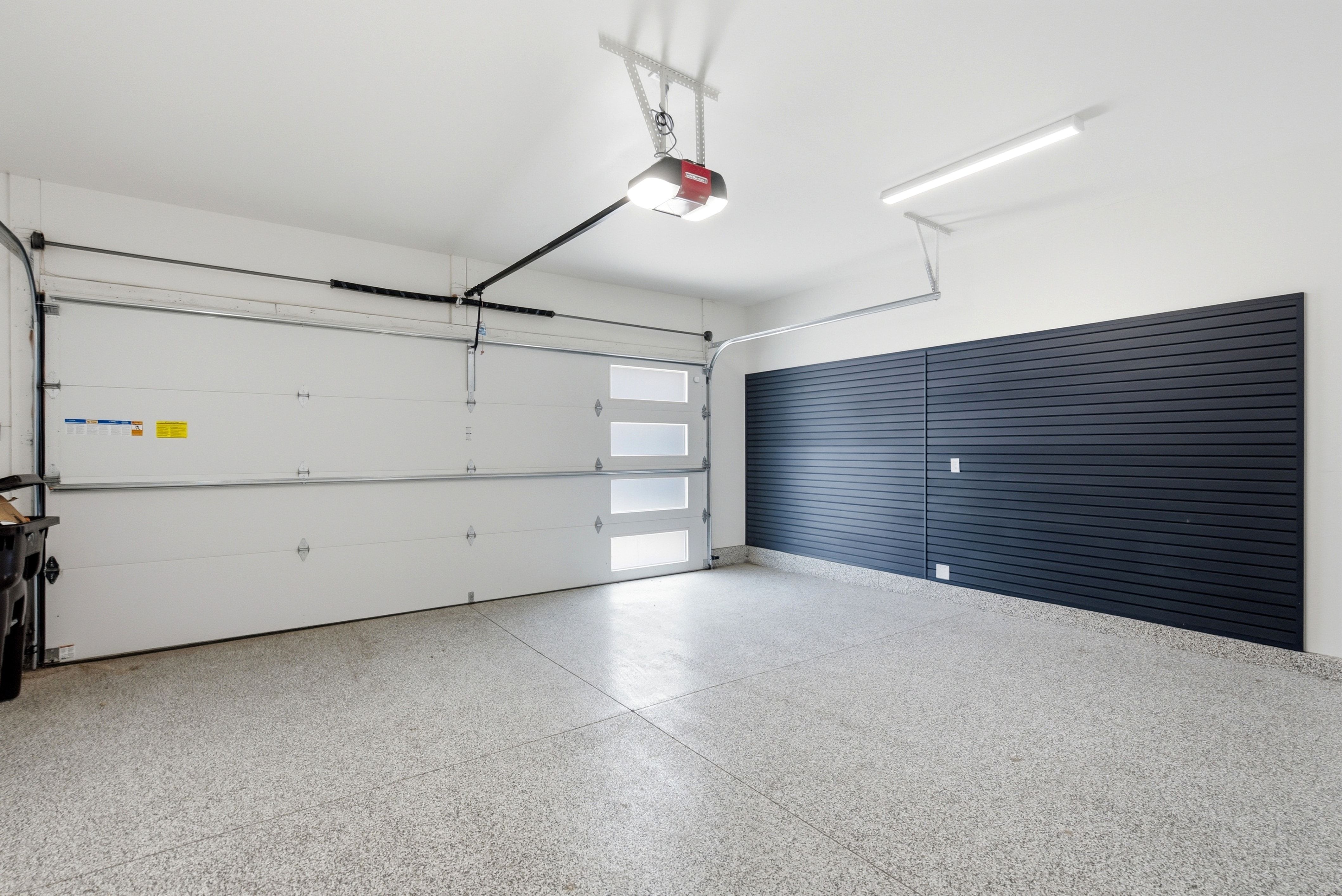 Attached two-car garage featuring a light gray epoxy floor coating, white overhead garage door with frosted glass panels, and a dark slatwall storage system
