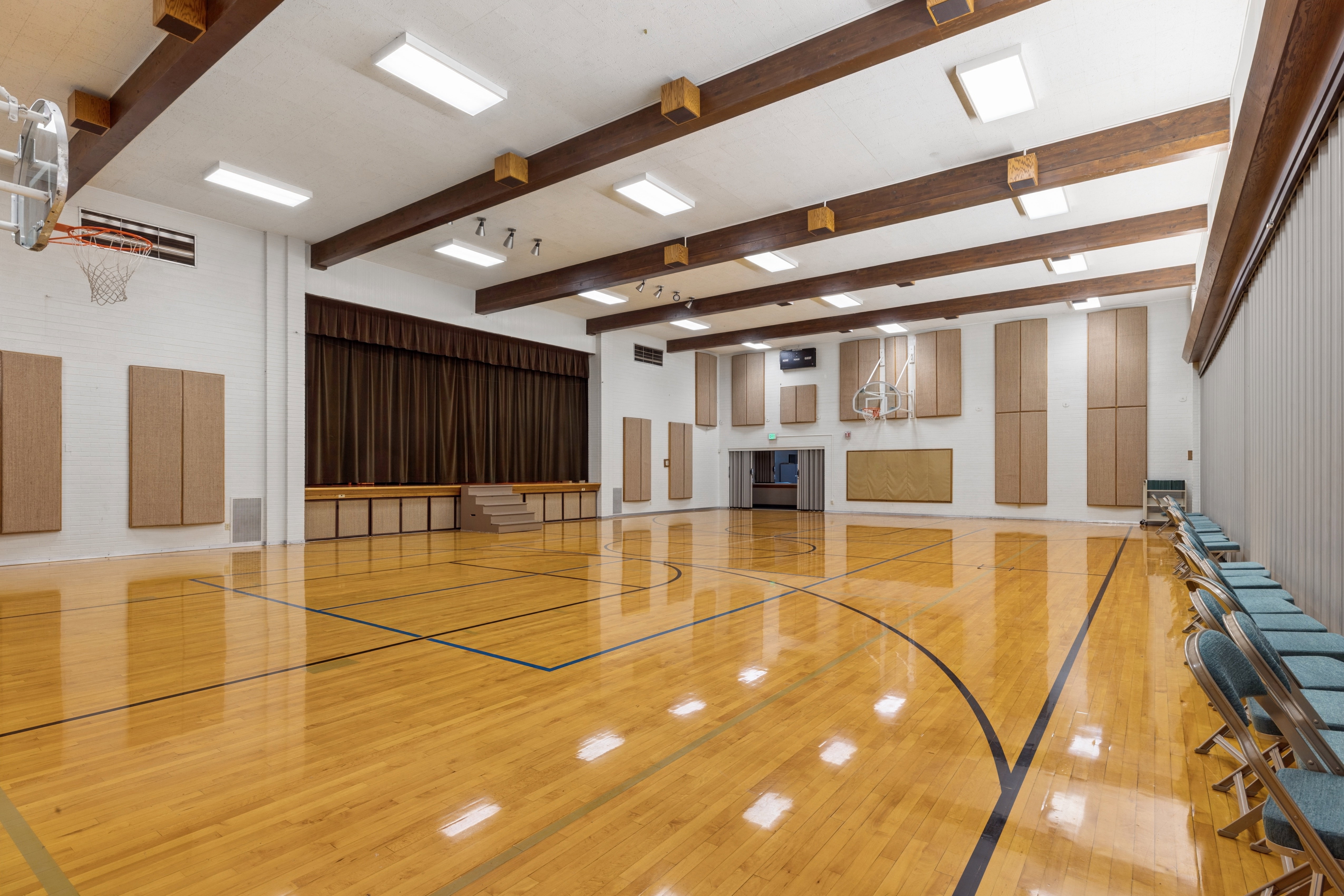 Versatile indoor court space featuring polished wood-finish flooring with court markings, exposed wood ceiling beams, and integrated stage with brown curtain backdrop