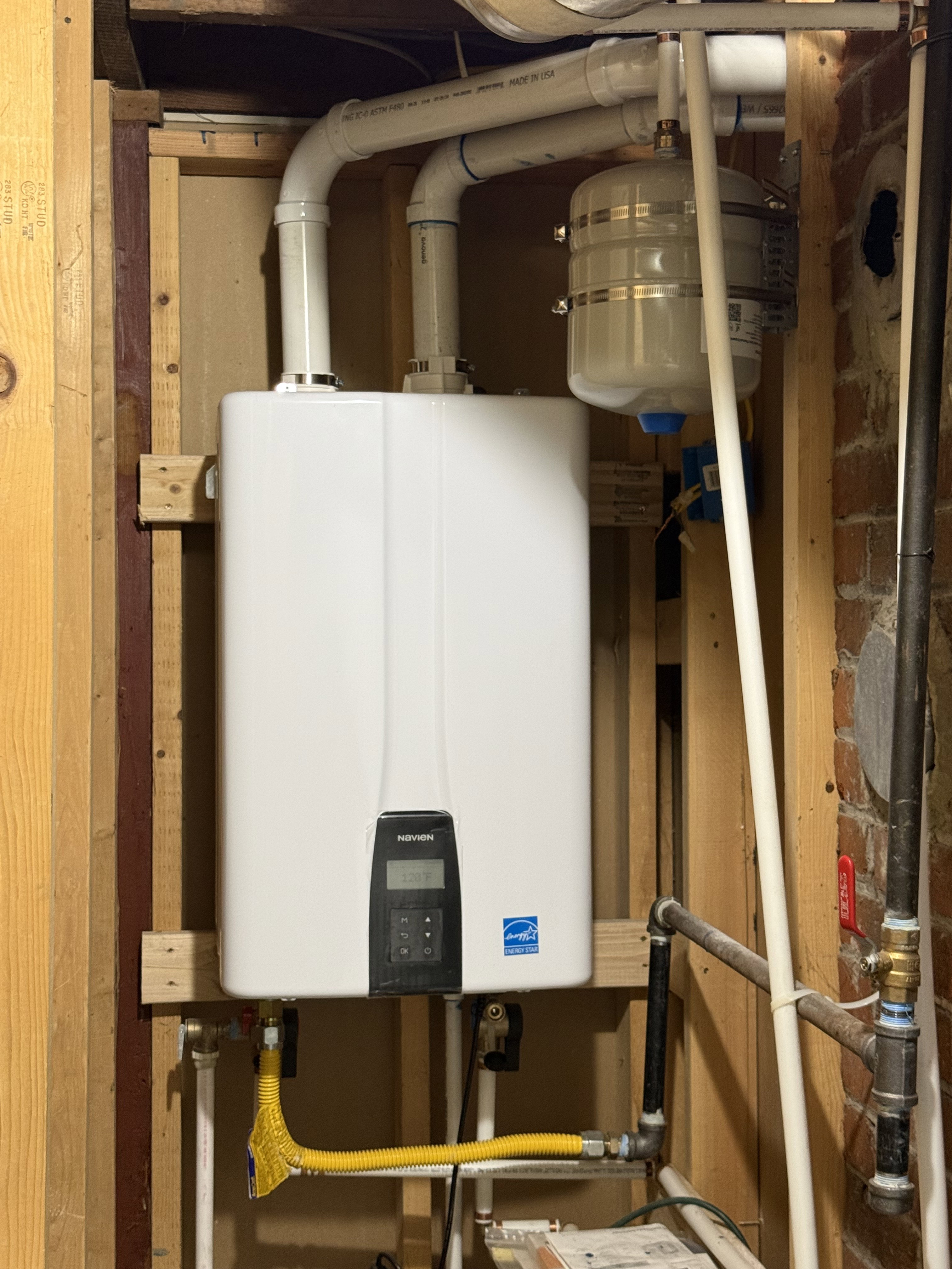 Wall-mounted tankless water heater with digital display, connected to PVC piping and a pressure expansion tank