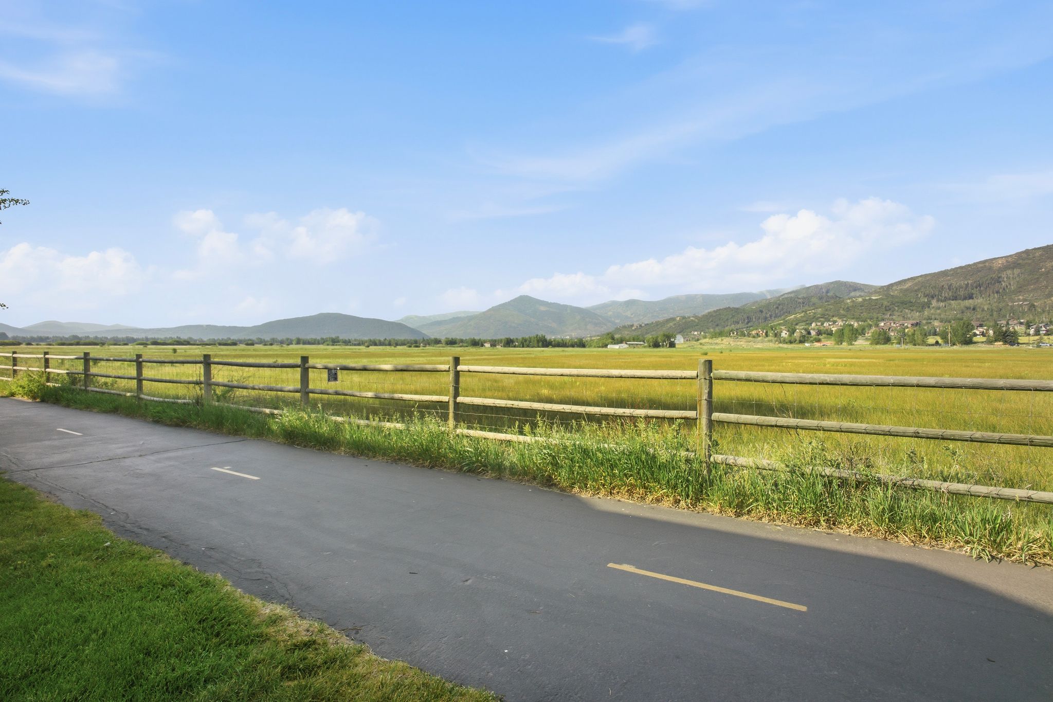Paved pathway bordered by a rustic wooden fence, alongside expansive green fields and distant mountains