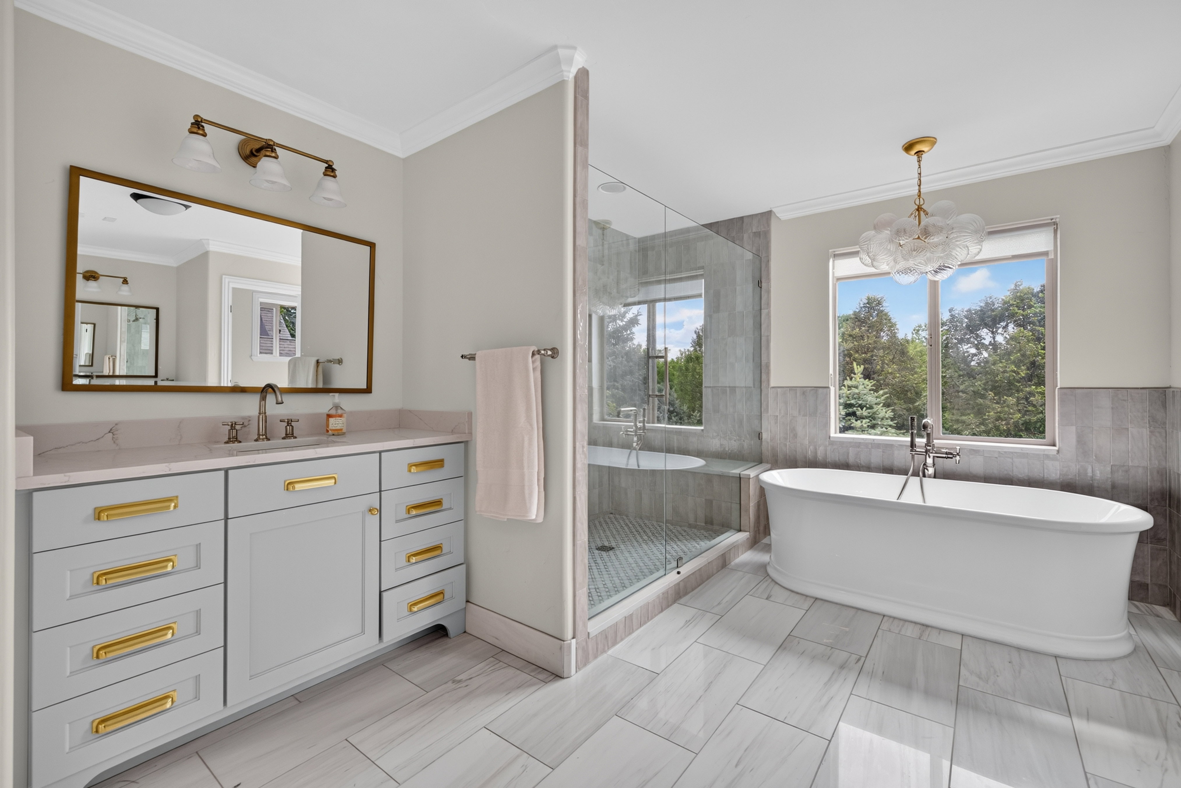 Bathroom featuring a freestanding tub, glass-enclosed shower, dual-sink vanity with gold hardware, large rectangular mirror, and ceiling-mounted chandelier