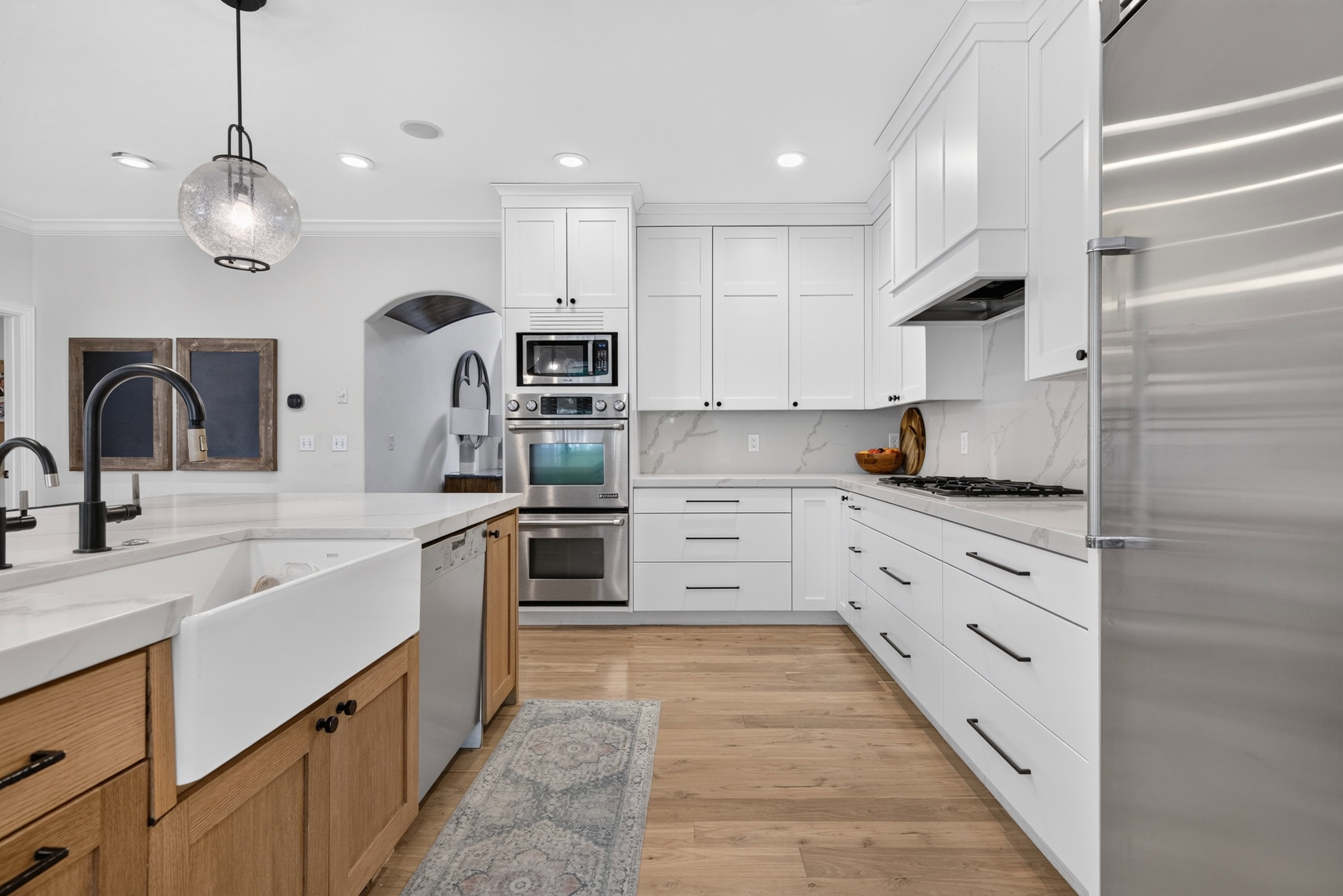 Gourmet kitchen featuring a farmhouse sink, stainless steel appliances, white shaker cabinetry, wood-finish flooring, and light-toned countertops with a matching backsplash