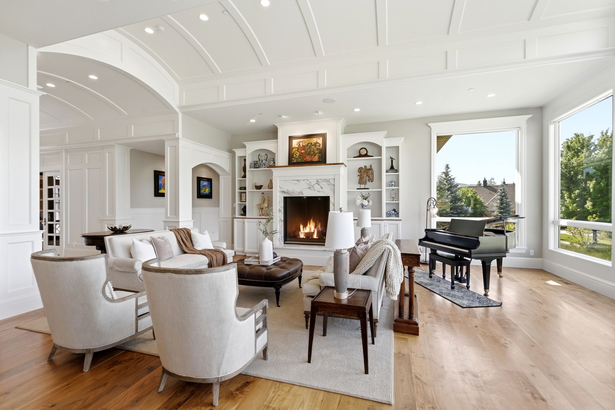 Spacious living area featuring light wood-finish flooring, a white mantel fireplace with a marble-style surround, built-in shelving, large picture windows, and a coffered ceiling