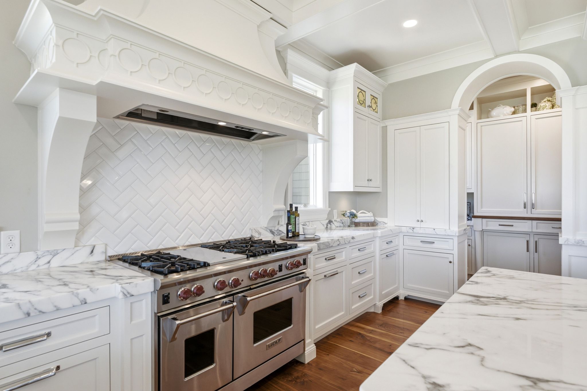 Gourmet kitchen featuring a professional-grade stainless steel range, custom white range hood with decorative molding, white subway tile backsplash in a herringbone pattern, white cabinetry with polished chrome hardware, and marble countertops