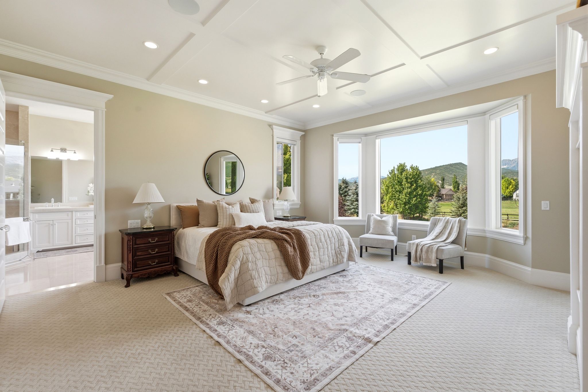 Spacious bedroom featuring a bay window, coffered ceiling with recessed lighting and ceiling fan, neutral carpet flooring, and an open doorway to a bathroom with a double vanity