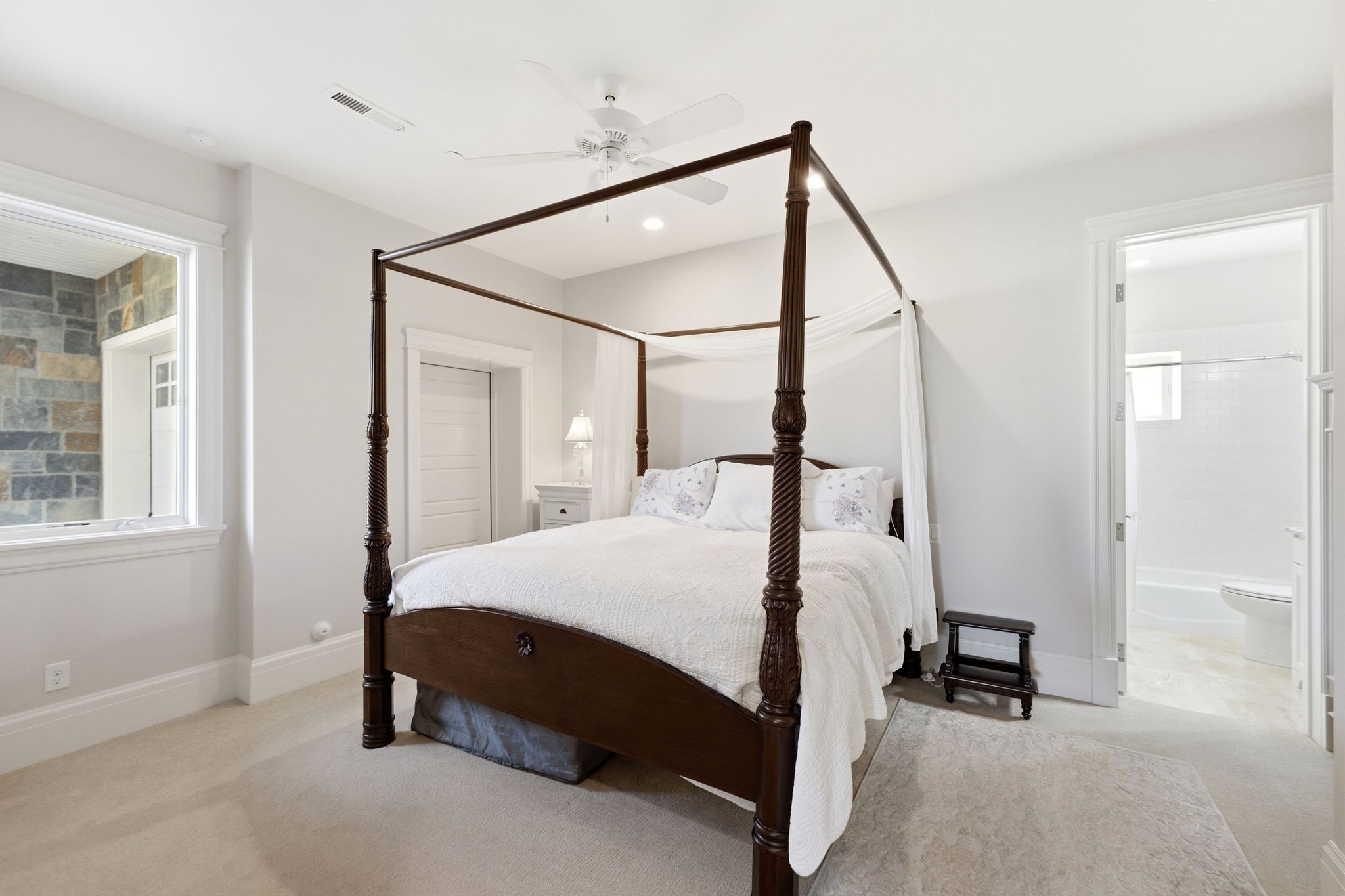 Carpeted bedroom featuring white walls, a ceiling fan, and a wide window with stone wall detailing