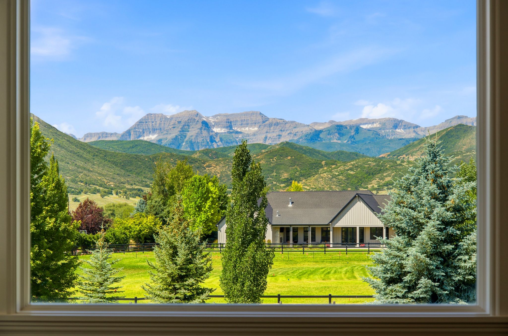 Window framing a distant residence with a dark roof and light-colored siding, surrounded by mature trees and expansive green grounds, set against a backdrop of mountains
