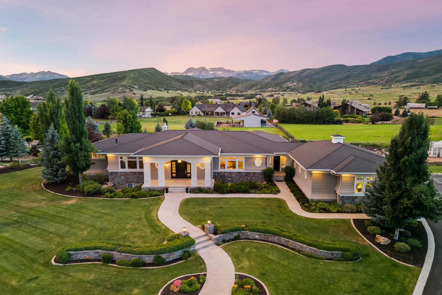Expansive property featuring a stone and siding exterior, a grand covered entry, a meticulously landscaped lawn, a winding paved pathway, and a distant mountainous backdrop