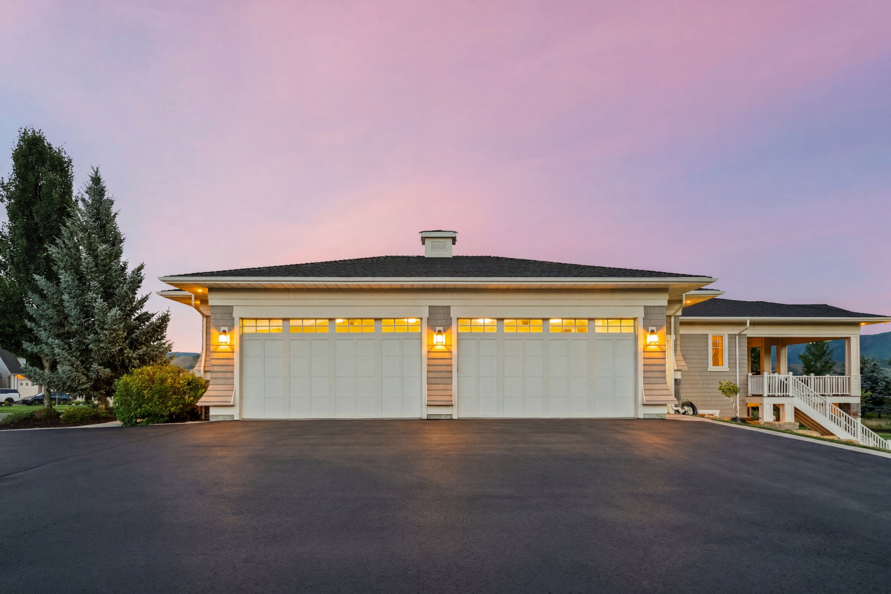 Exterior featuring a large asphalt driveway, two garage doors with upper window panels, and wall-mounted exterior light fixtures