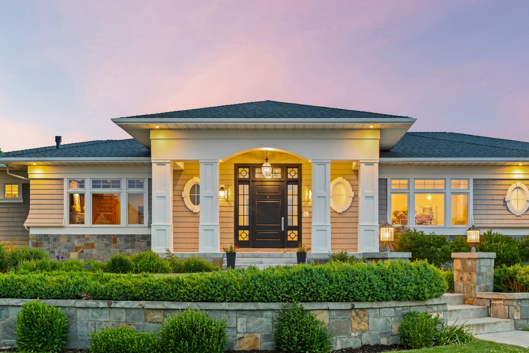Inviting facade featuring a grand portico entry, horizontal siding, stone veneer accents, multiple window styles including oval and casement, and manicured shrubbery with stone retaining walls