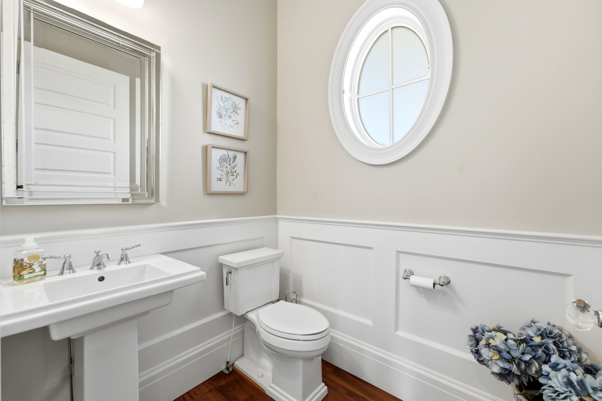 Bathroom featuring wainscoting, an oval window, a pedestal sink with a polished chrome faucet, a rectangular mirror, and wood-finish flooring