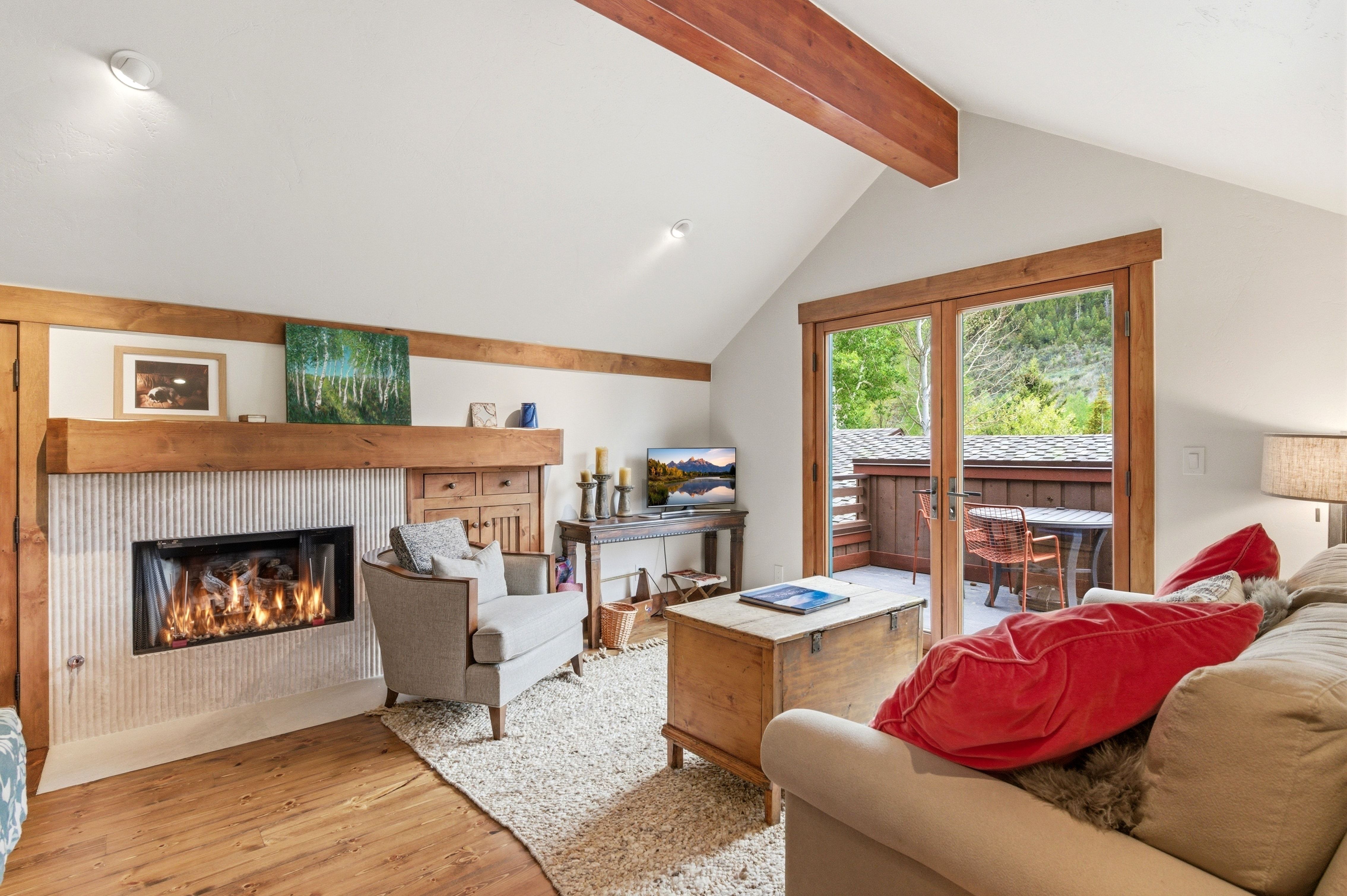 Vaulted ceiling living space featuring exposed wood beams, a modern fireplace with a textured surround, wood-finish flooring, and direct access to an outdoor patio through double glass doors