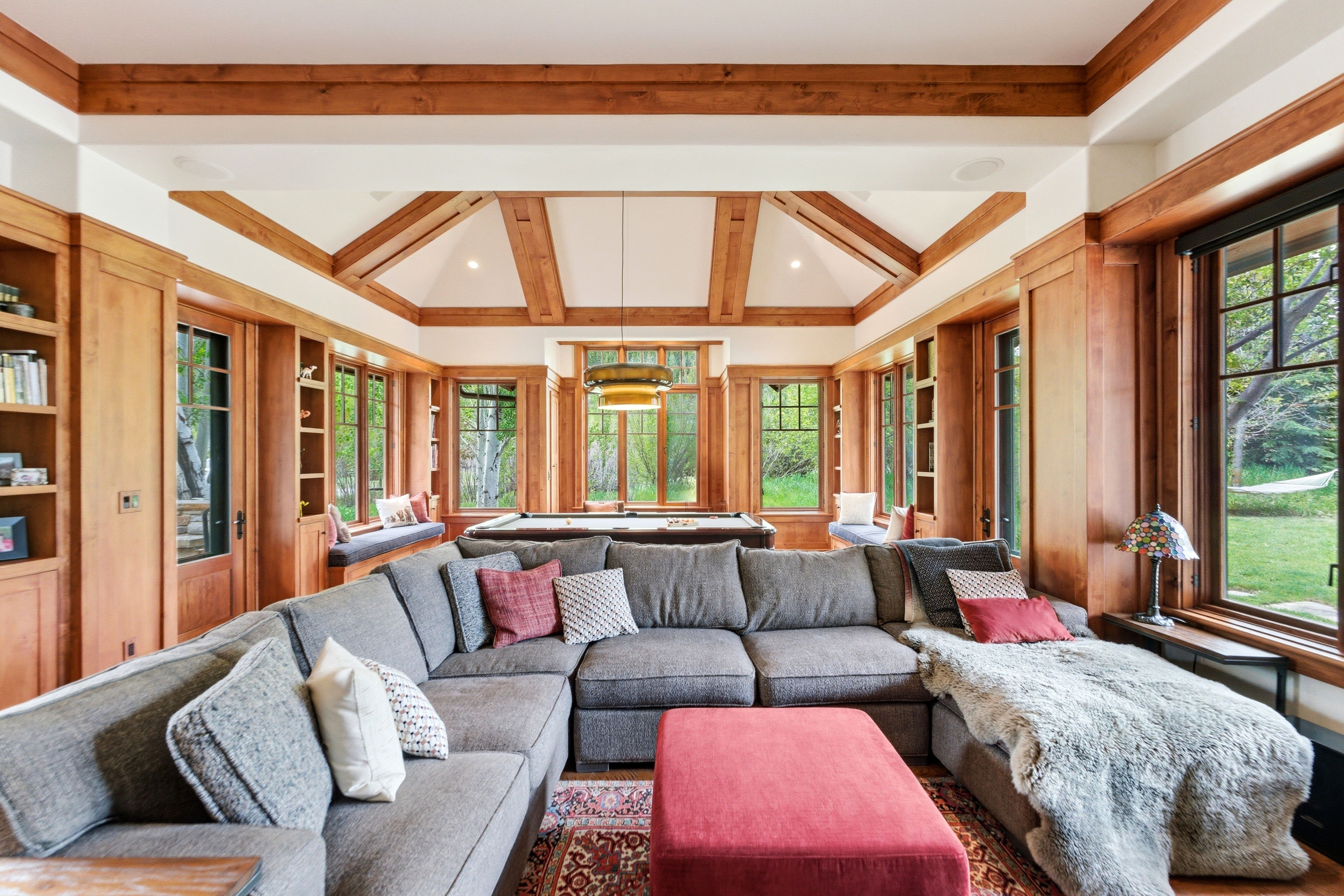 Spacious recreation room featuring exposed wood beam ceilings, extensive built-in wood cabinetry, multiple large windows, and window seats