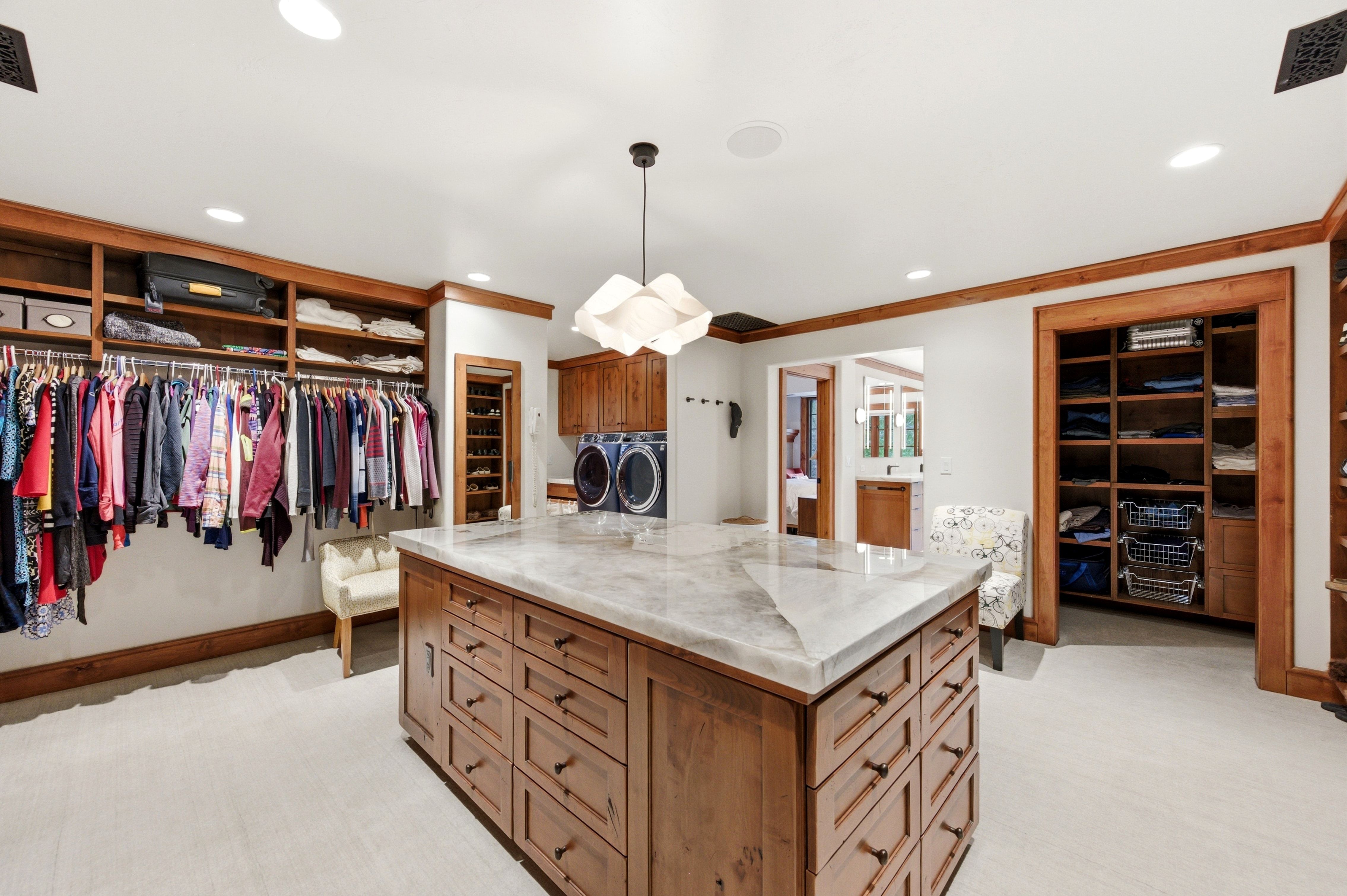 Expansive custom closet featuring a large central island with a stone countertop and abundant drawer storage, integrated laundry appliances, extensive built-in shelving, and warm wood trim