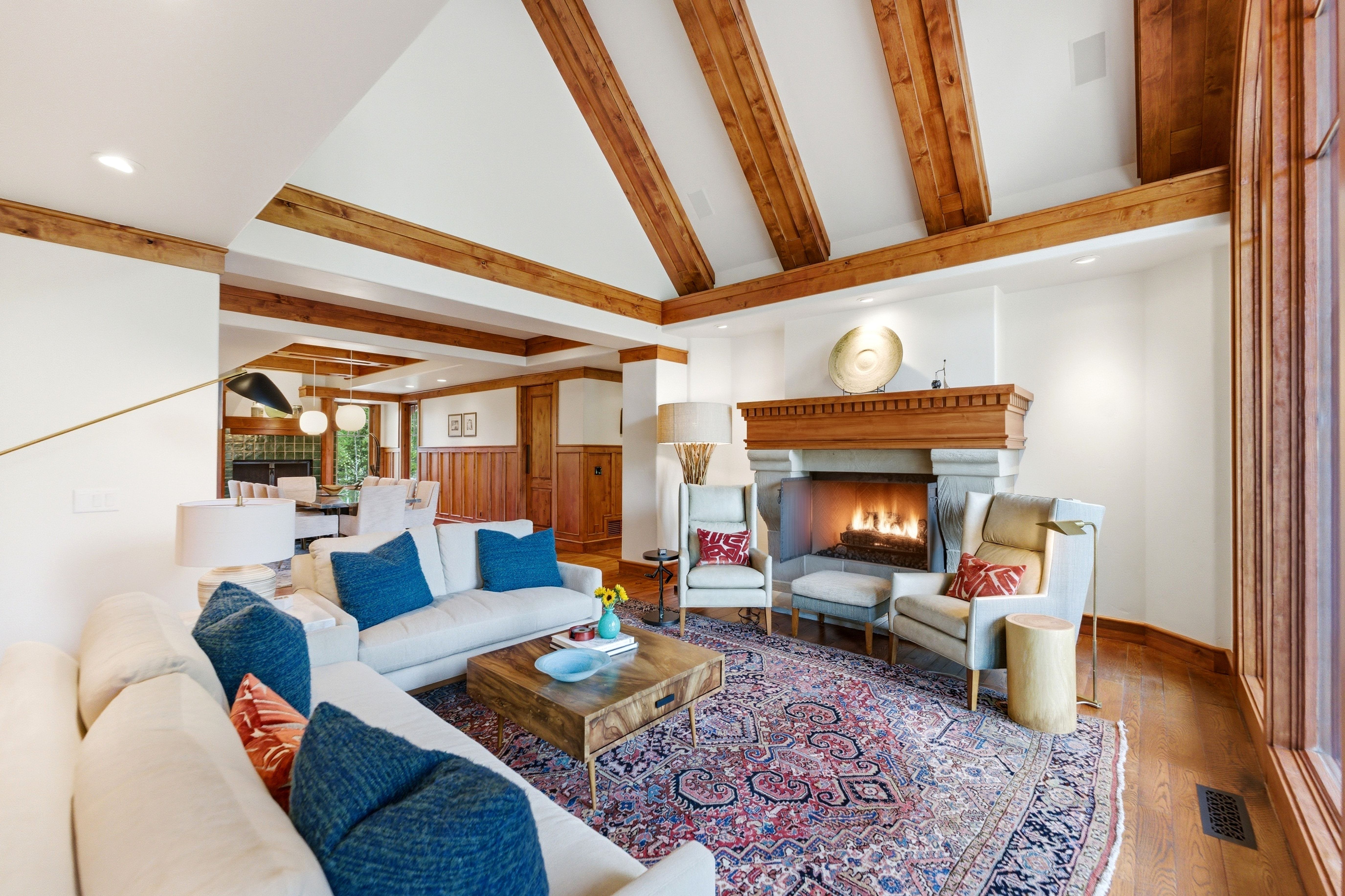 Living space featuring a stone-clad fireplace with wood mantel, exposed ceiling beams, wood-finish flooring, and expansive windows