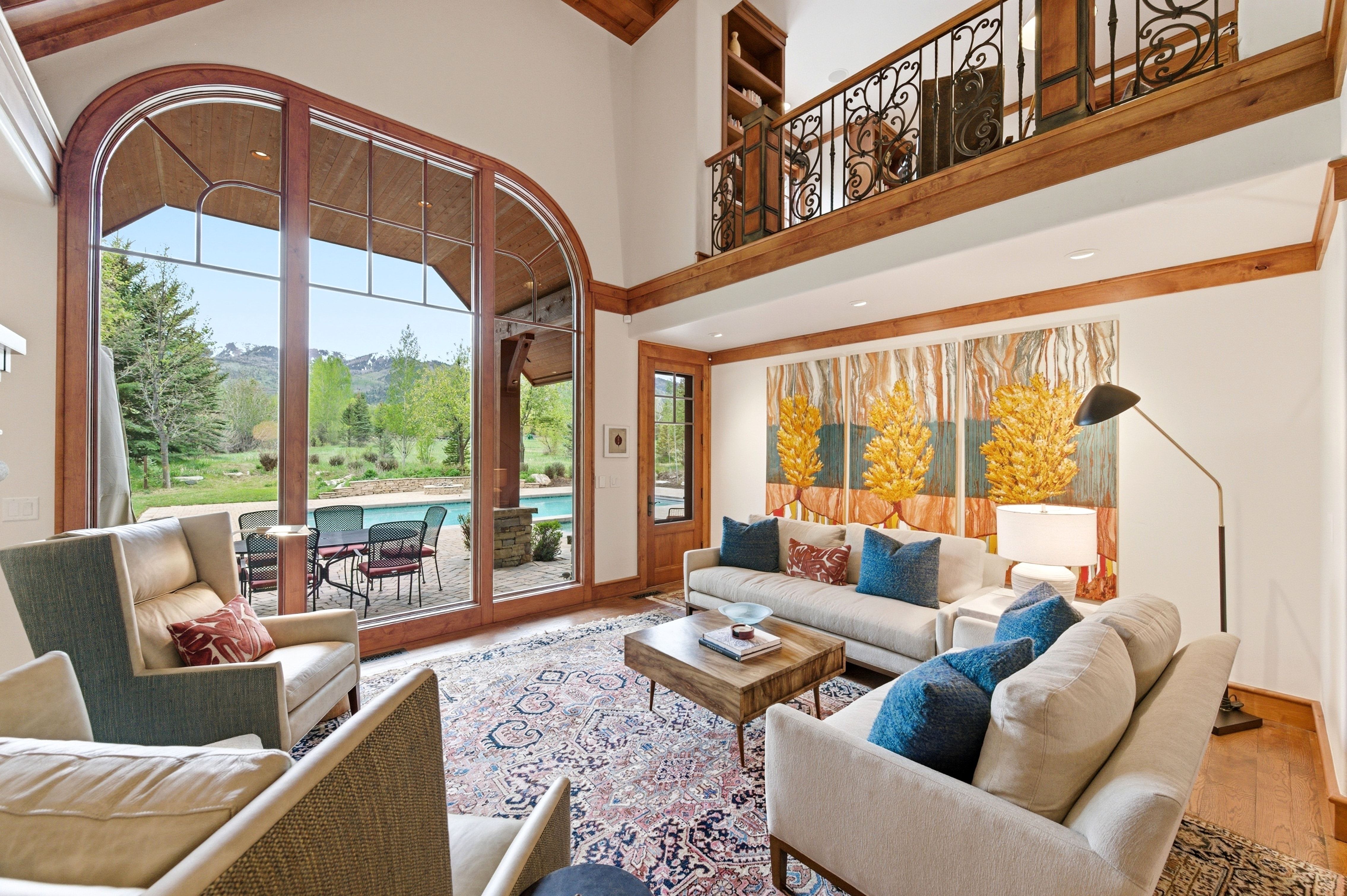Spacious living area featuring a two-story arched window wall with wood framing, exposed wood beams, and a decorative wrought iron balcony overlooking the space