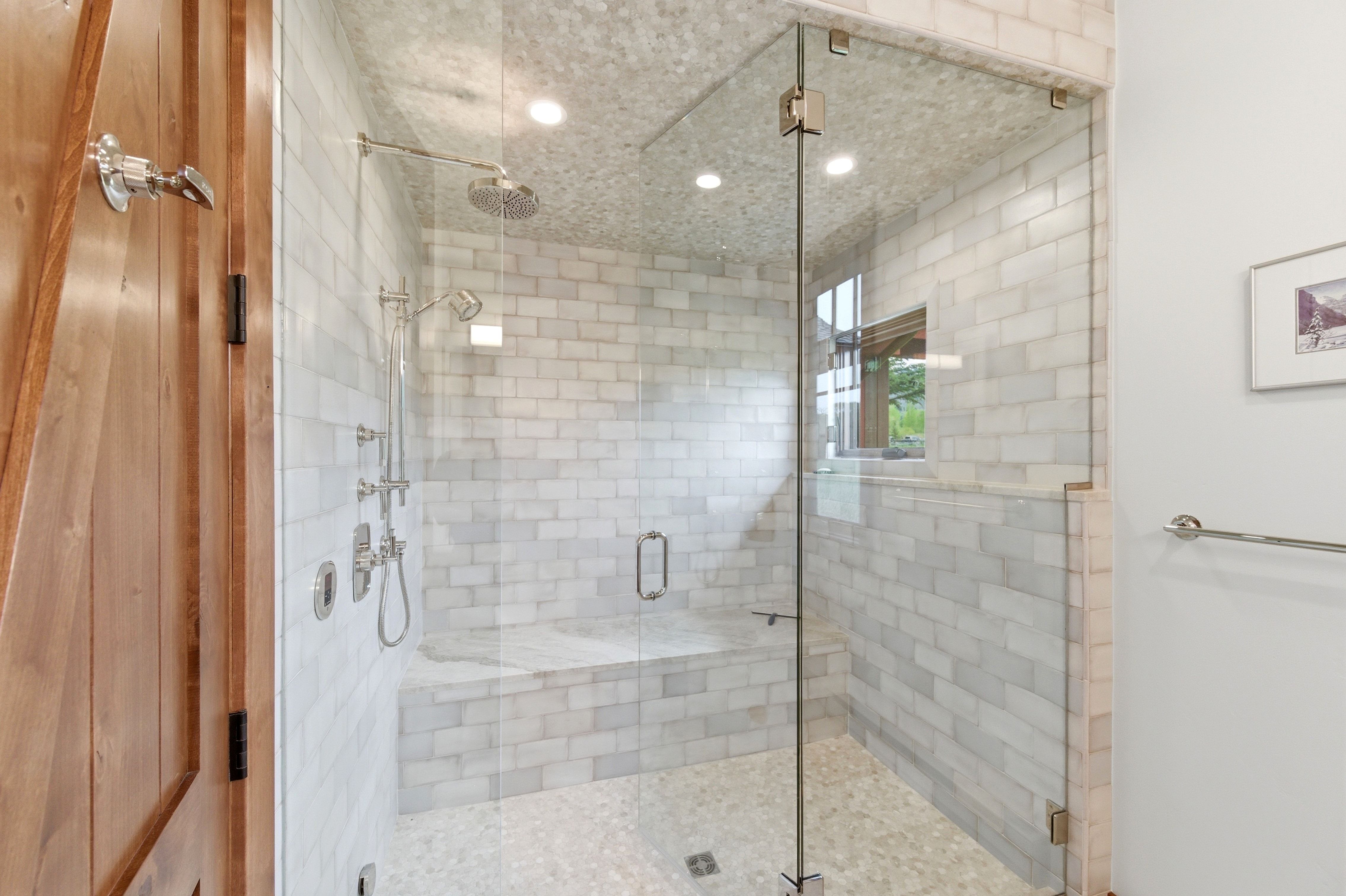 Spacious shower enclosure with glass surround, subway tile walls, pebble tile flooring, built-in bench, and a rain shower head with additional handheld sprayer
