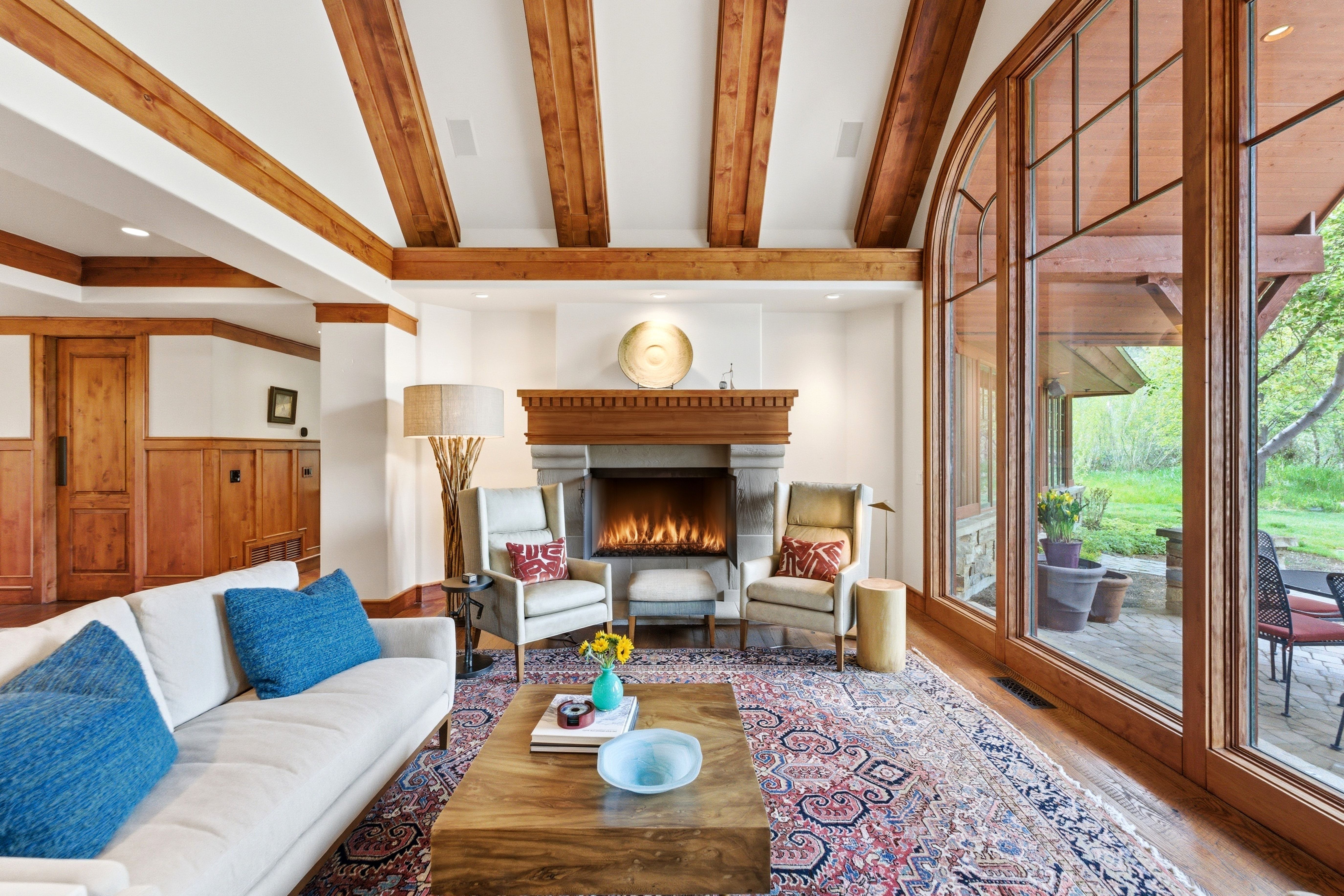 Living area featuring wood-beam ceilings, a stone fireplace with a wood mantel, expansive arched windows, hardwood flooring, and wood paneling