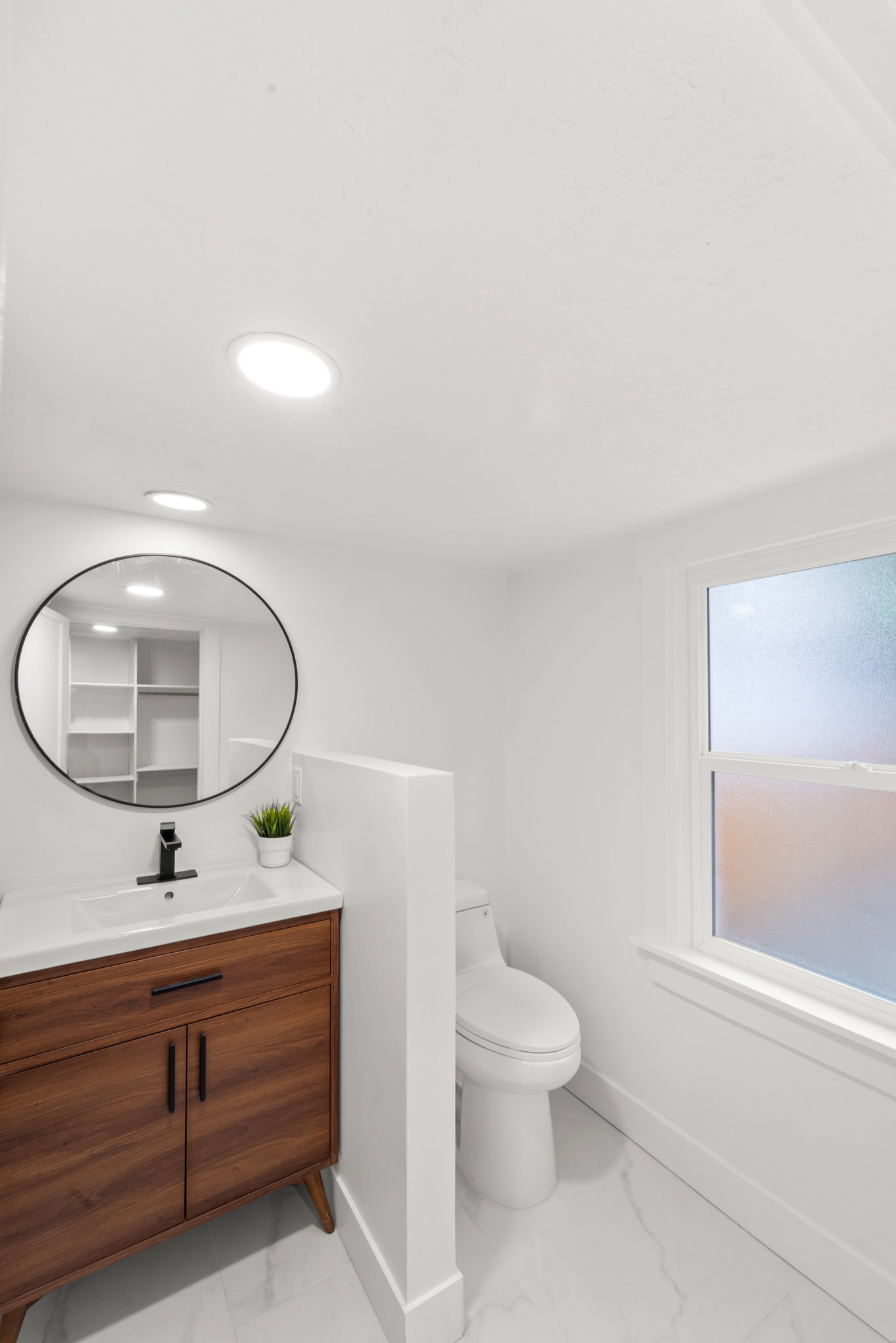 Primary full bathroom featuring a wood-finish vanity with integrated sink, a circular black-framed mirror, recessed lighting, and white marble-look floor tiles