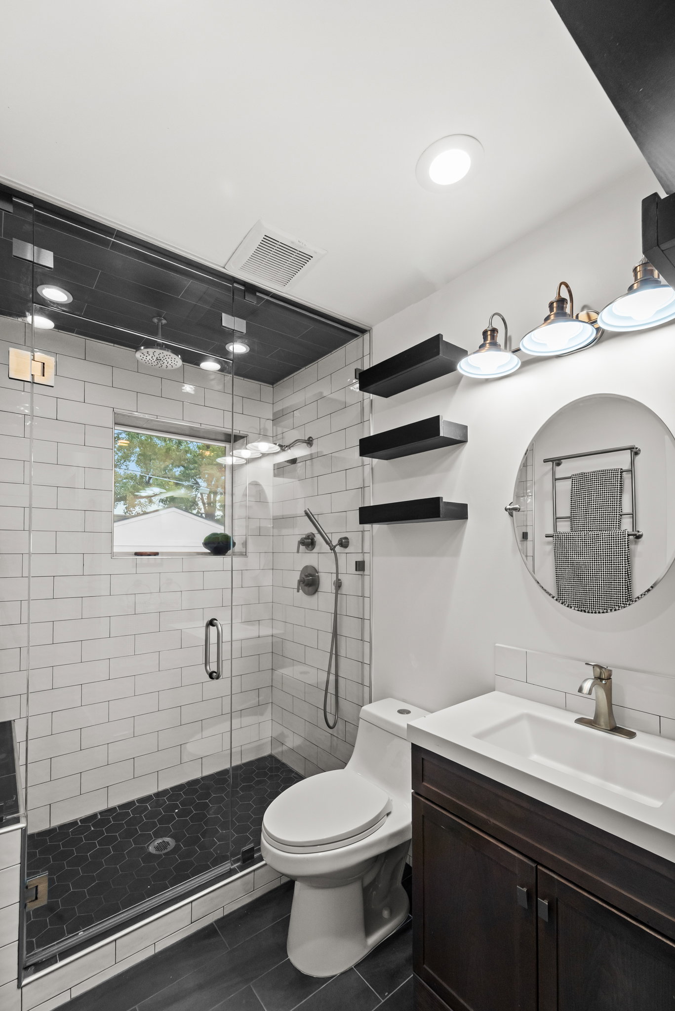 Contemporary bathroom featuring a dark wood-finish vanity with a white integrated sink, a framed glass shower enclosure with white subway tile walls, a dark hexagonal tile shower floor, and dark wood-finish flooring
