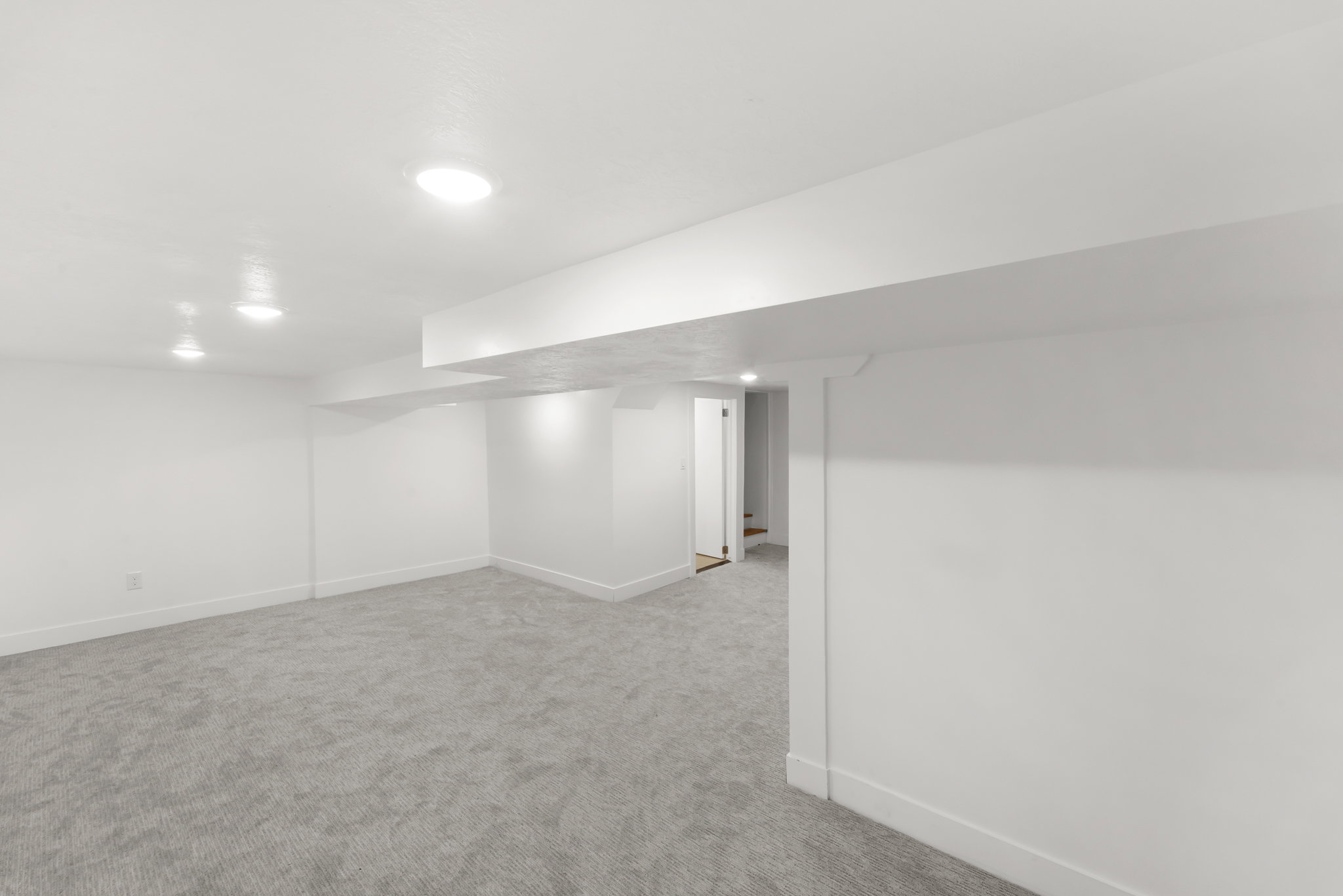 Spacious room featuring light gray carpet, recessed lighting, white walls, and a visible doorway leading to a staircase
