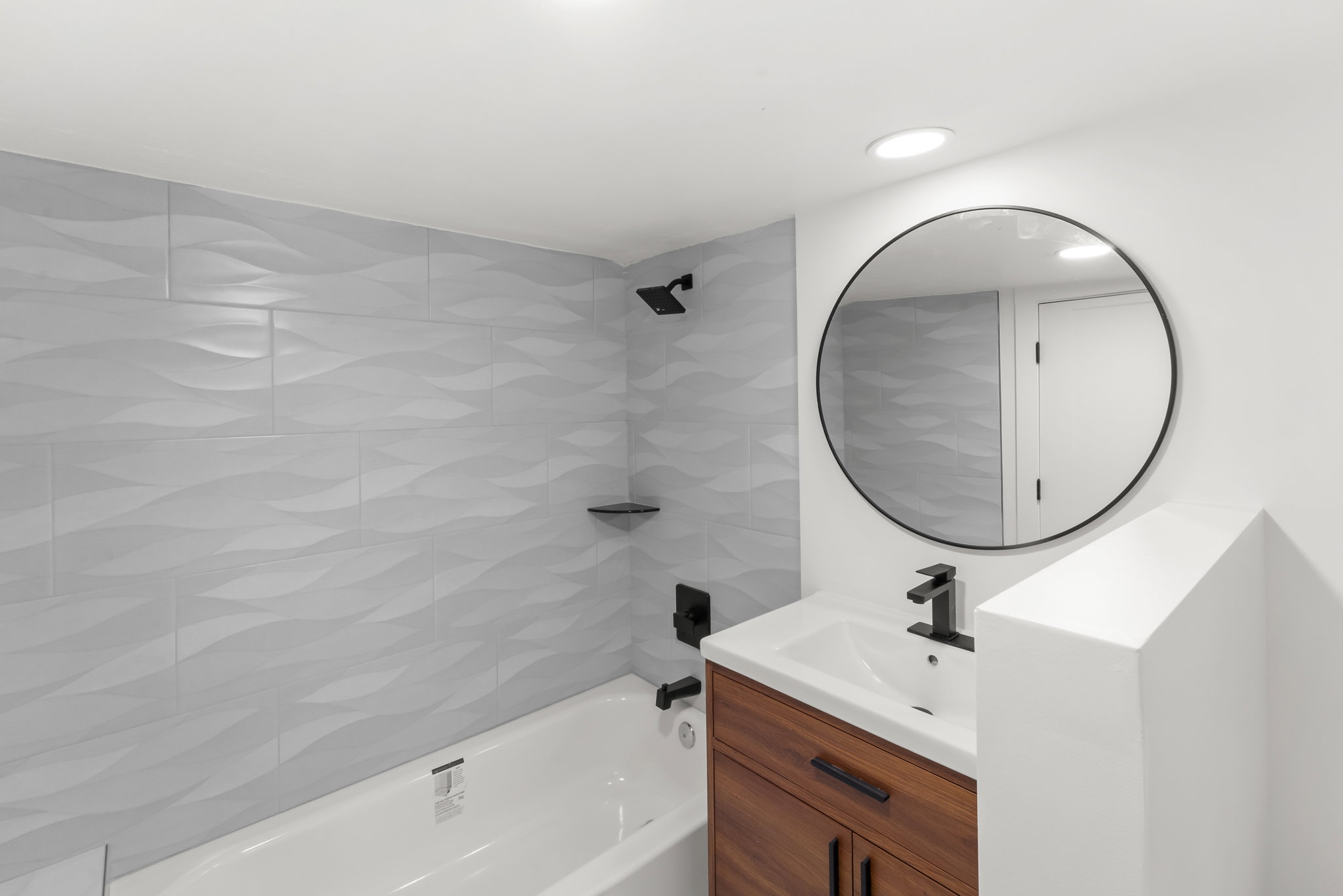 Primary full bathroom featuring a white bathtub with light gray textured tile surround, a dark wood-finish vanity with a white countertop sink, matte black faucet, and a round black-framed mirror