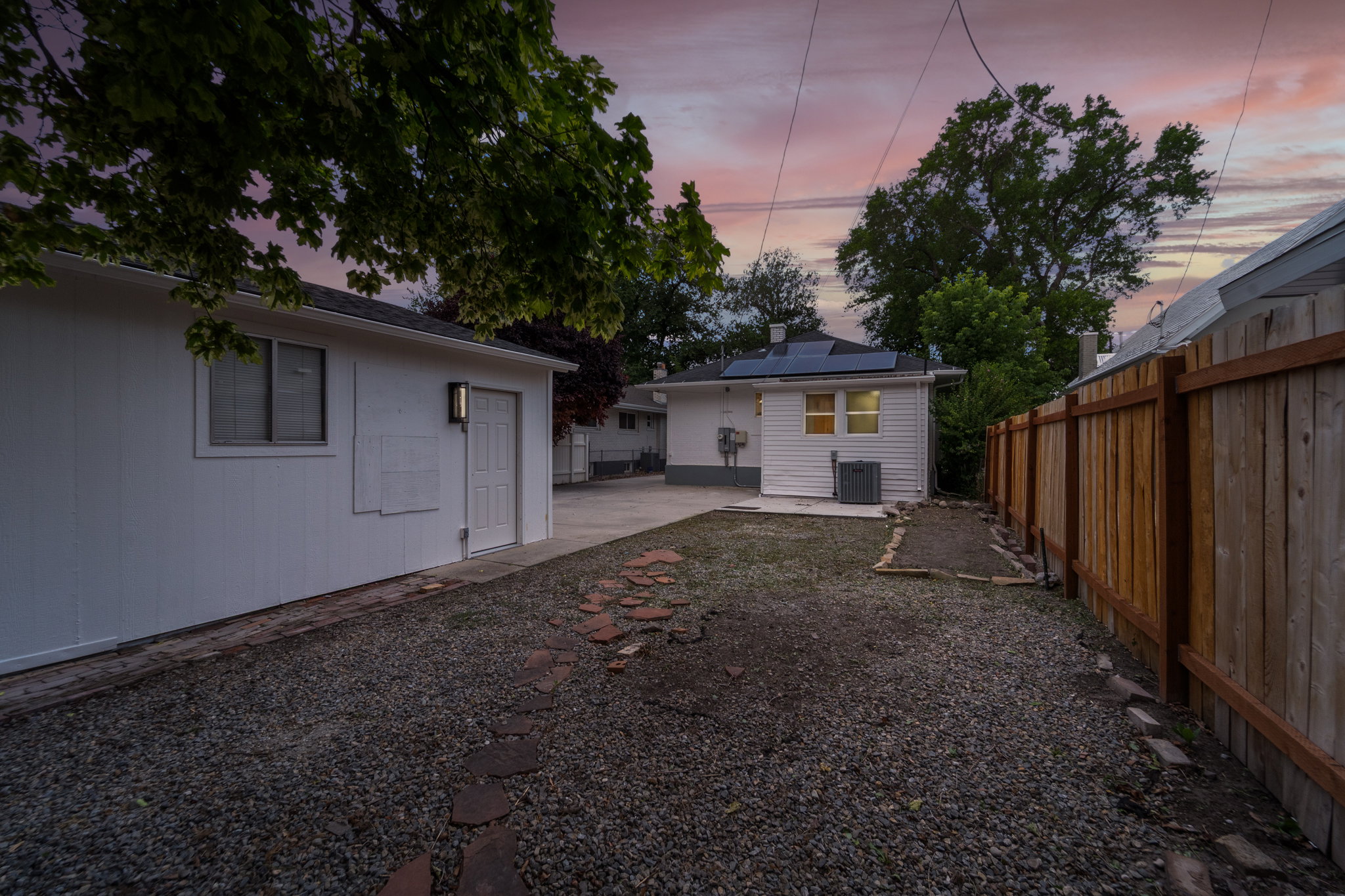 Expansive gravel yard featuring a wood privacy fence, concrete patio, and a separate white accessory structure with solar panels