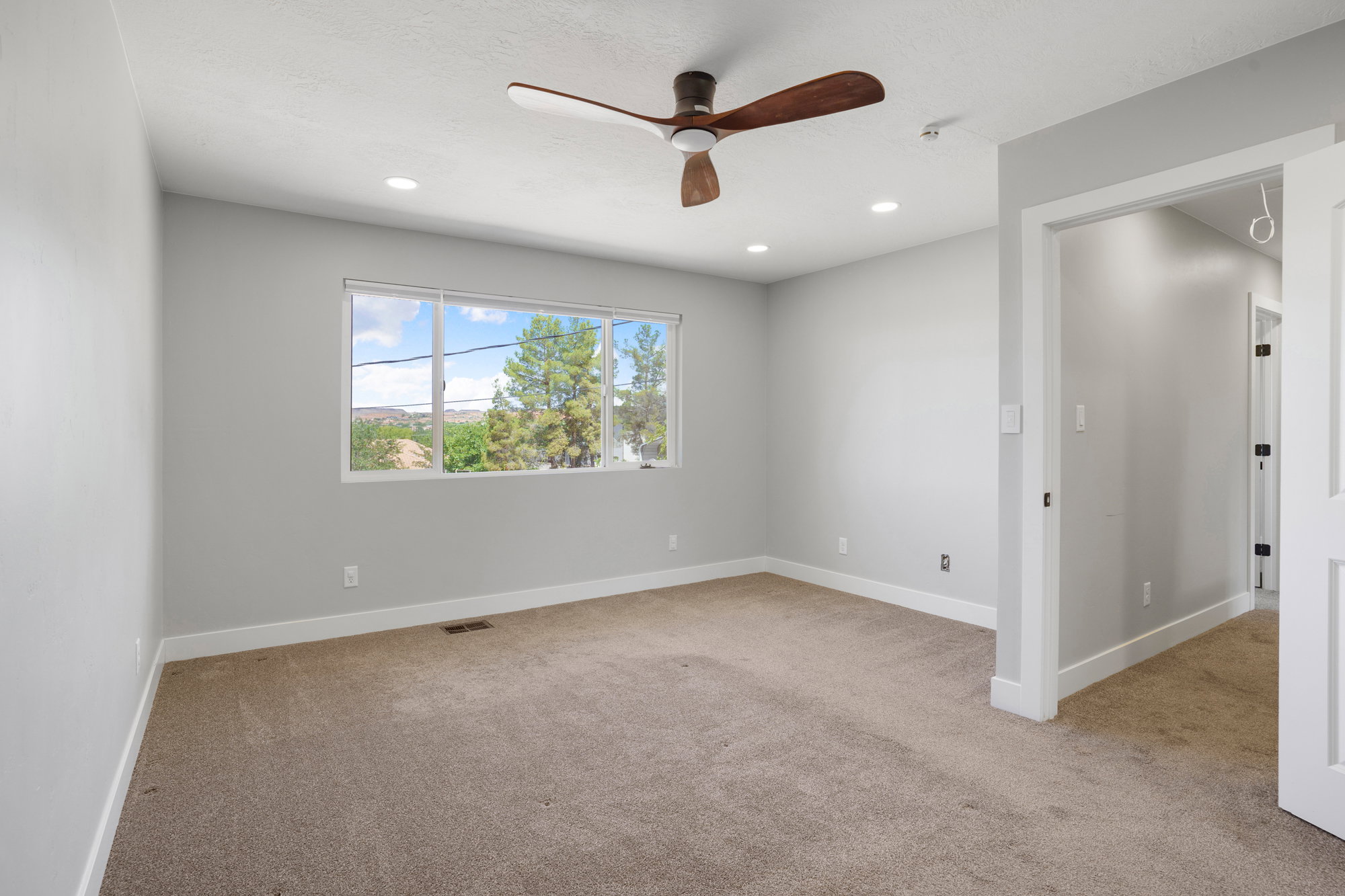 Room featuring light gray walls, brown carpeting, a large multi-pane window, and a wood-finish ceiling fan with integrated lighting