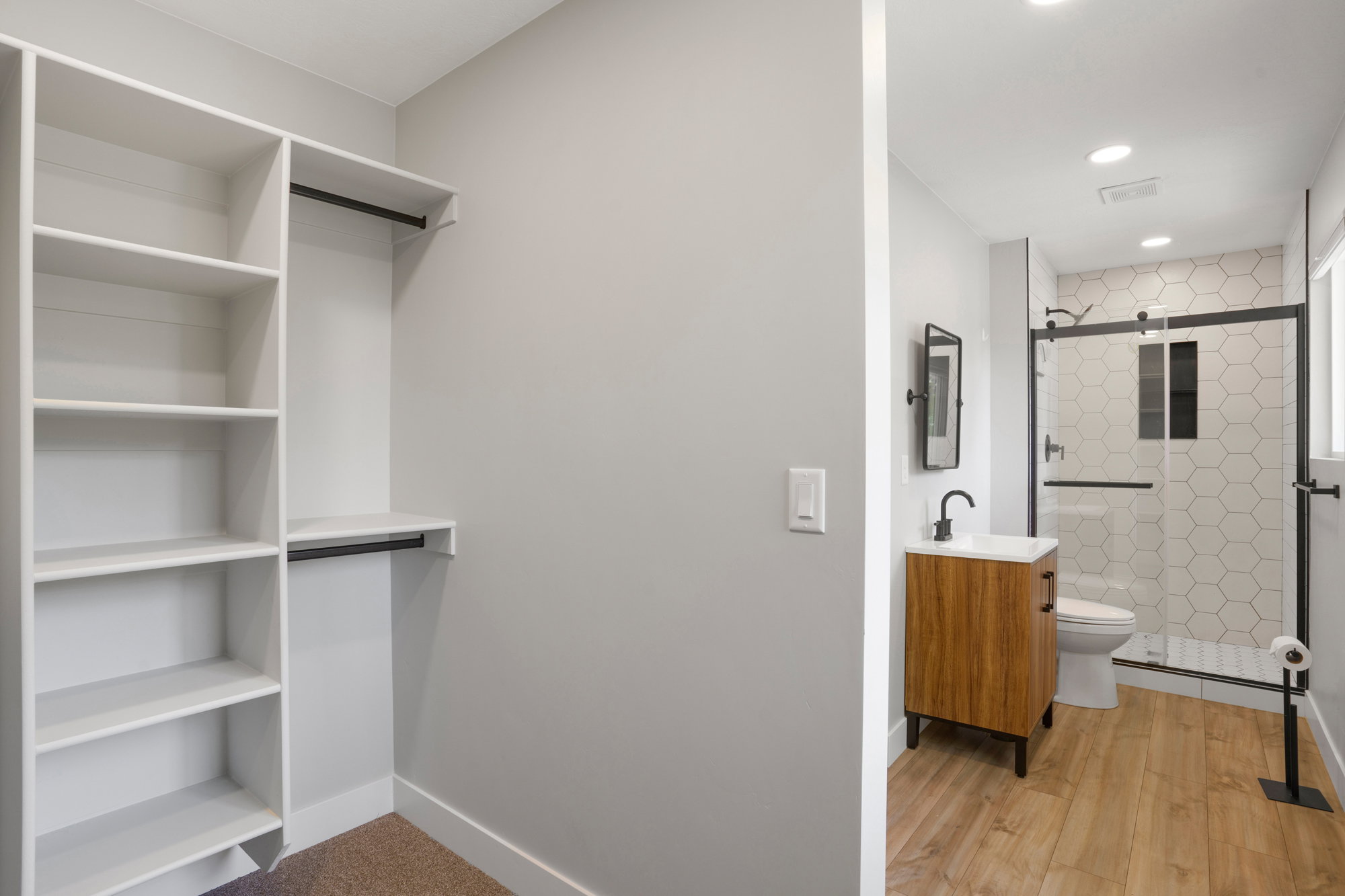 Built-in closet shelving and hanging rods with a bathroom featuring a wood-finish vanity, matte black fixtures, a walk-in shower with hexagonal tile, and wood-finish flooring