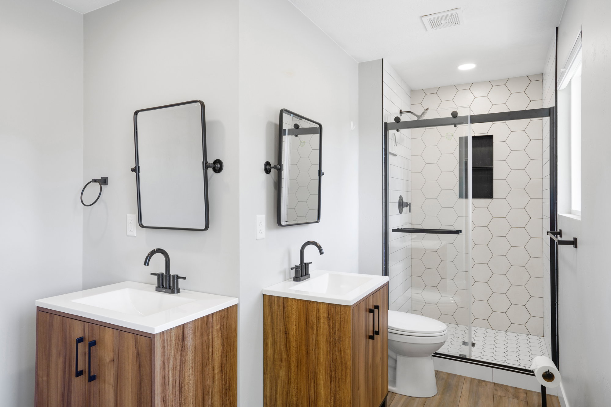 Contemporary bathroom featuring a wood-finish vanity with a white countertop and integrated sink, matte black fixtures, a shower with a sliding glass door and hexagonal tile surround, and wood-finish flooring