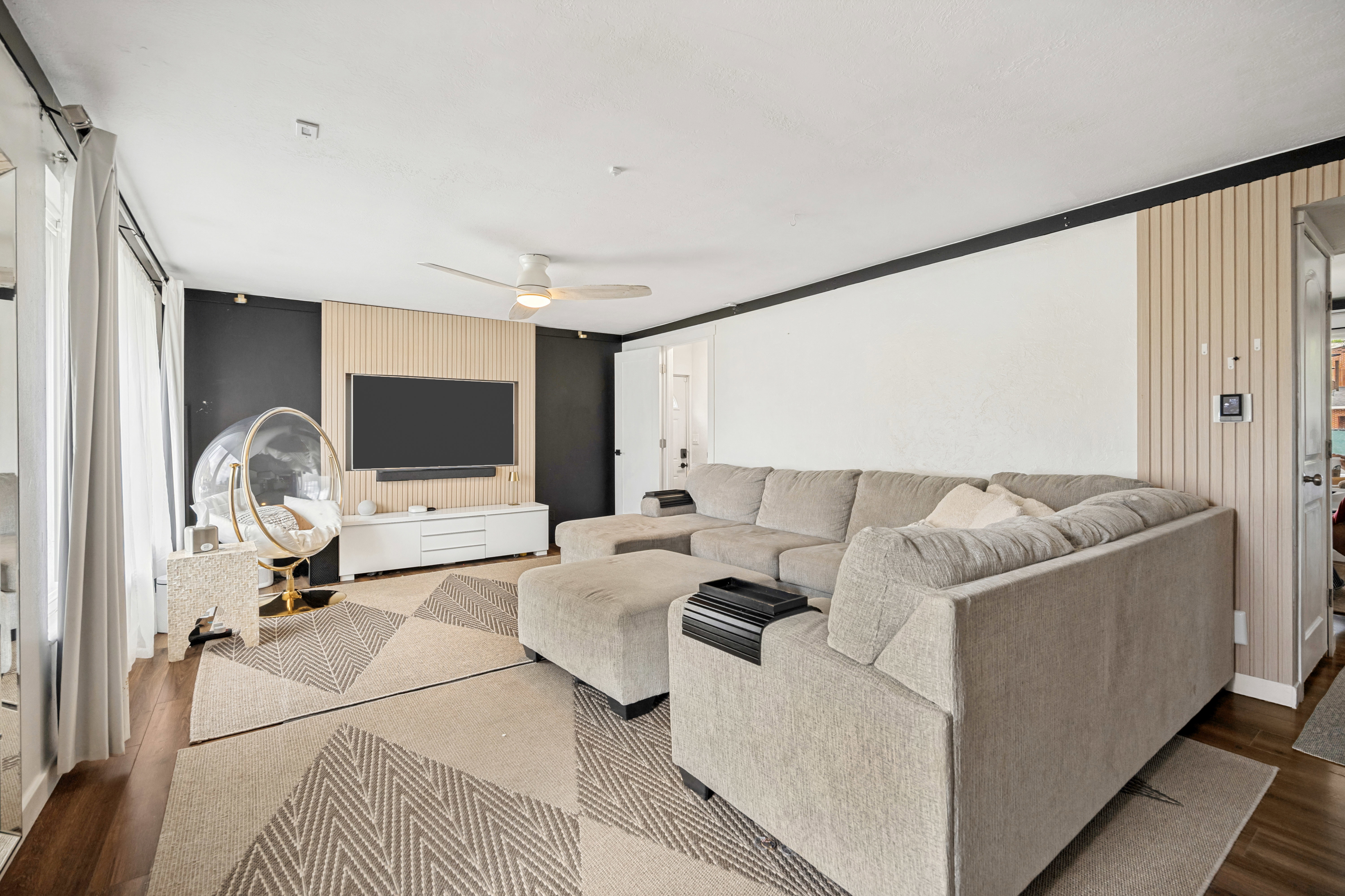 Living area featuring wood-finish flooring, a contemporary accent wall with vertical slat paneling, and a ceiling fan with integrated lighting