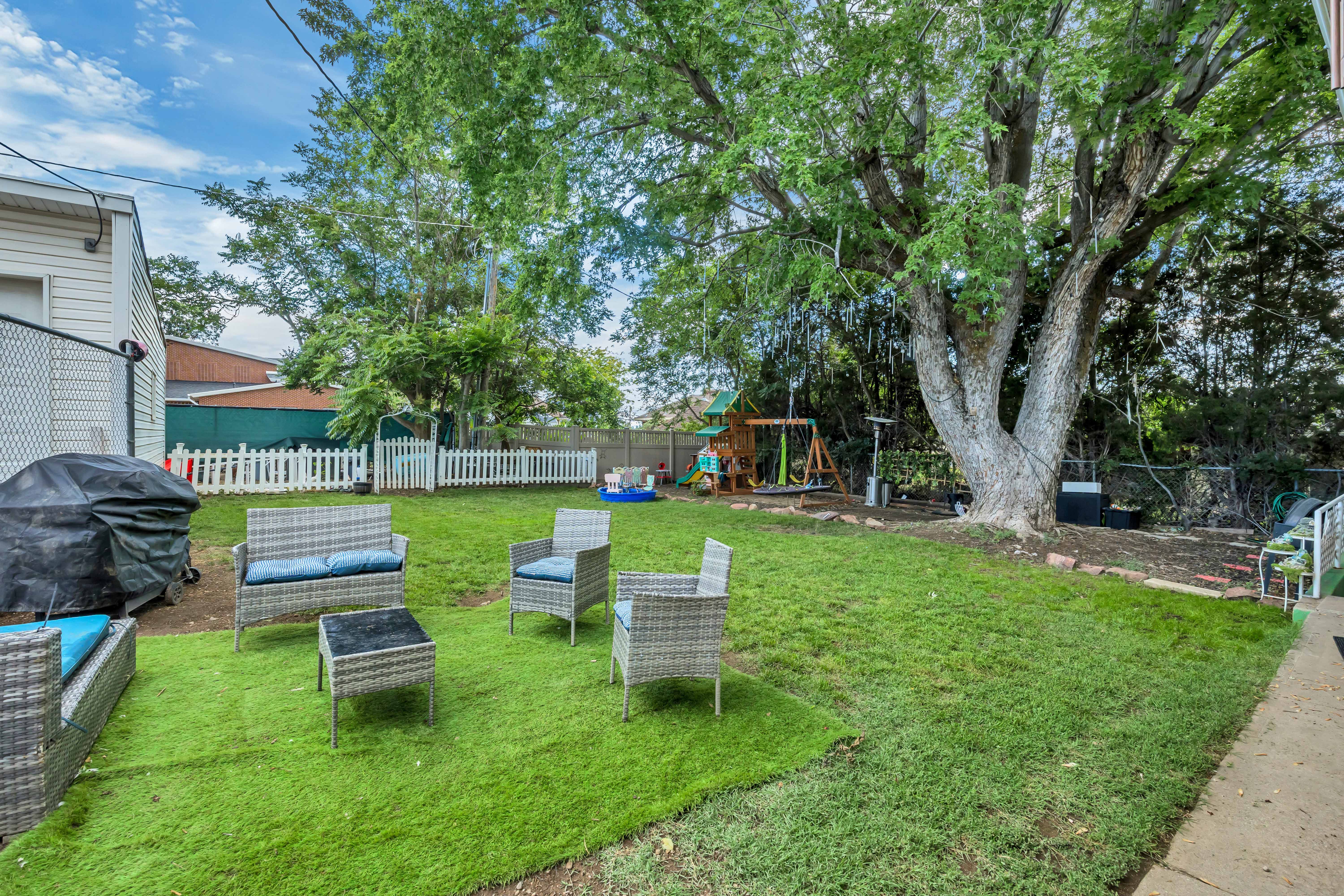 Expansive backyard featuring a large tree, green lawn, white picket fence, and a patio area with artificial turf
