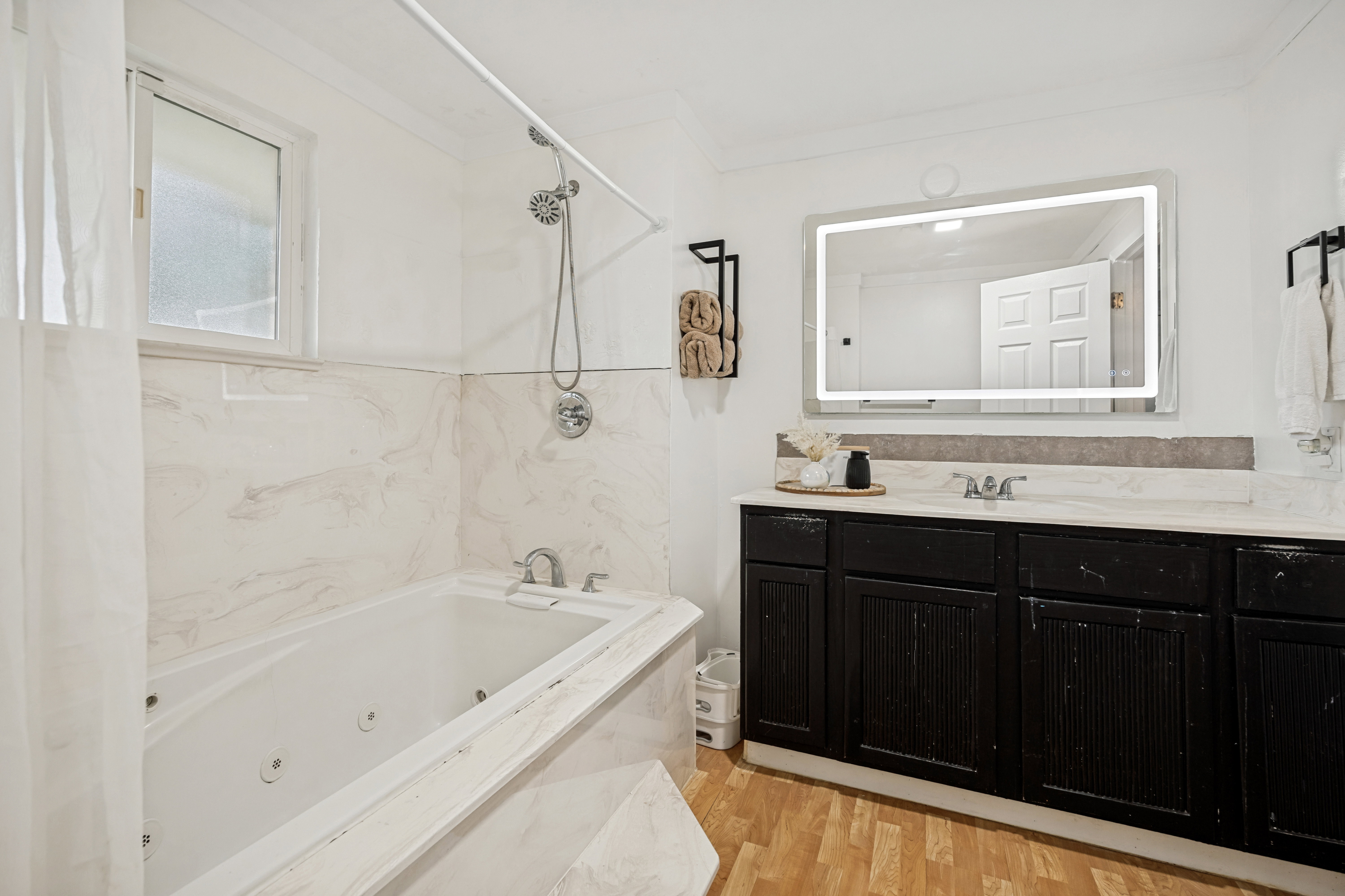 Bathroom featuring a jetted tub with marble-look surround, a black vanity with marble-look countertop, a back-lit vanity mirror, and wood-finish flooring