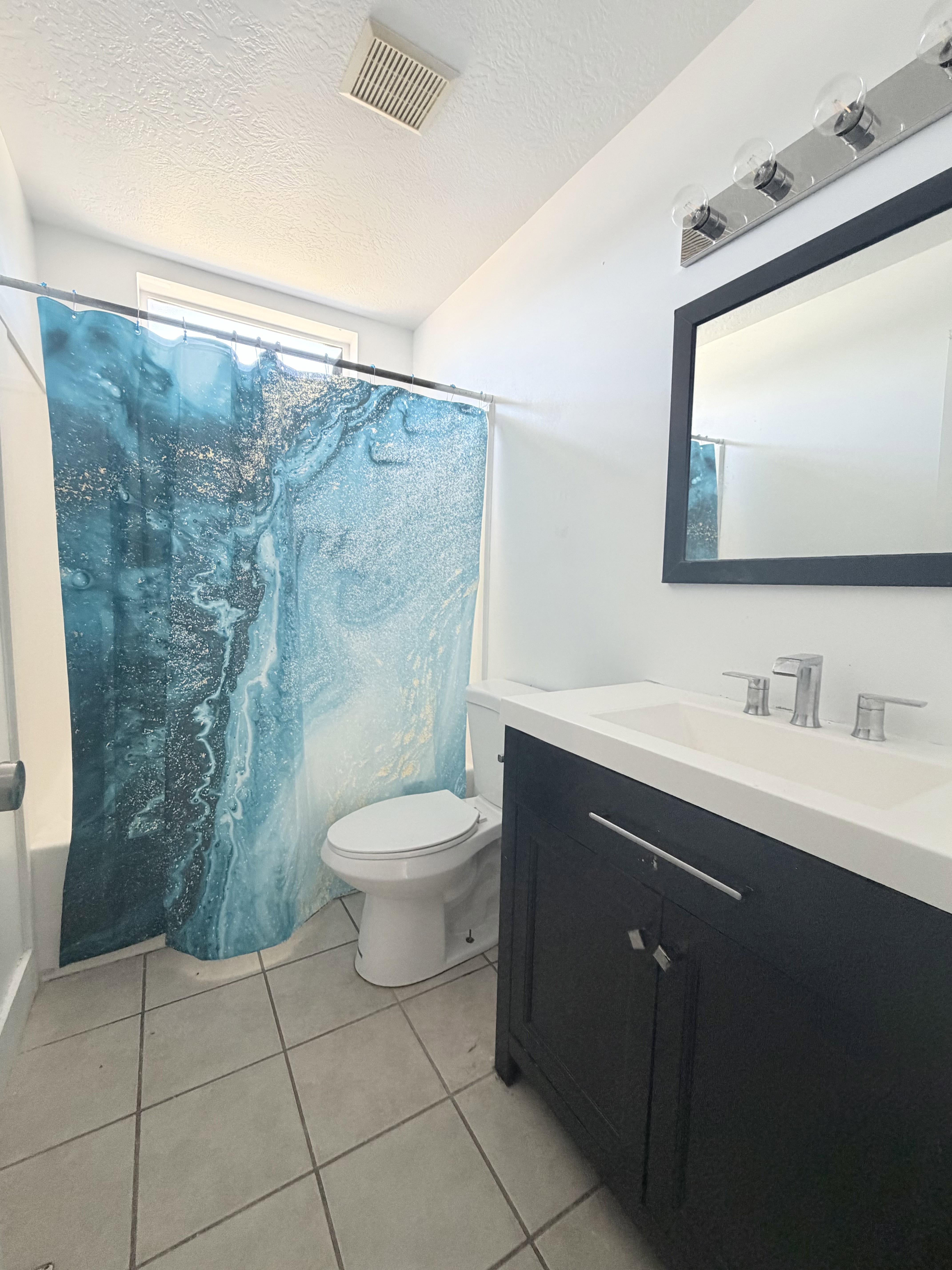 Bathroom featuring a black vanity with a white countertop and integrated sink, chrome faucet, wall-mounted mirror, and tile flooring