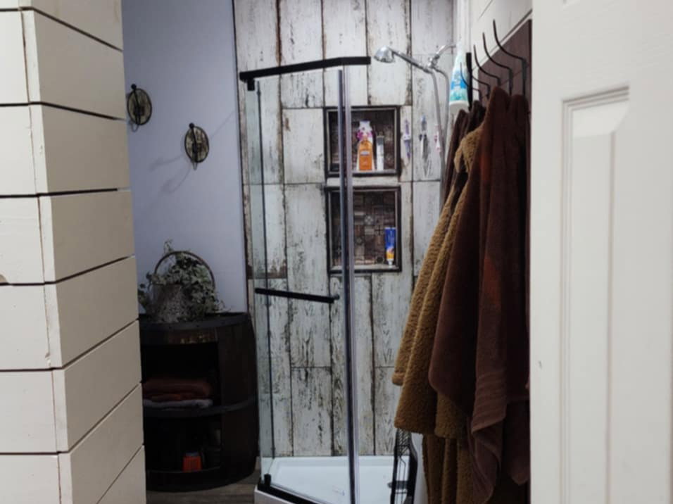Hallway entrance featuring a prominent wood beam, shiplap wall paneling, and wood-finish flooring leading to a bathroom with a glass-enclosed shower