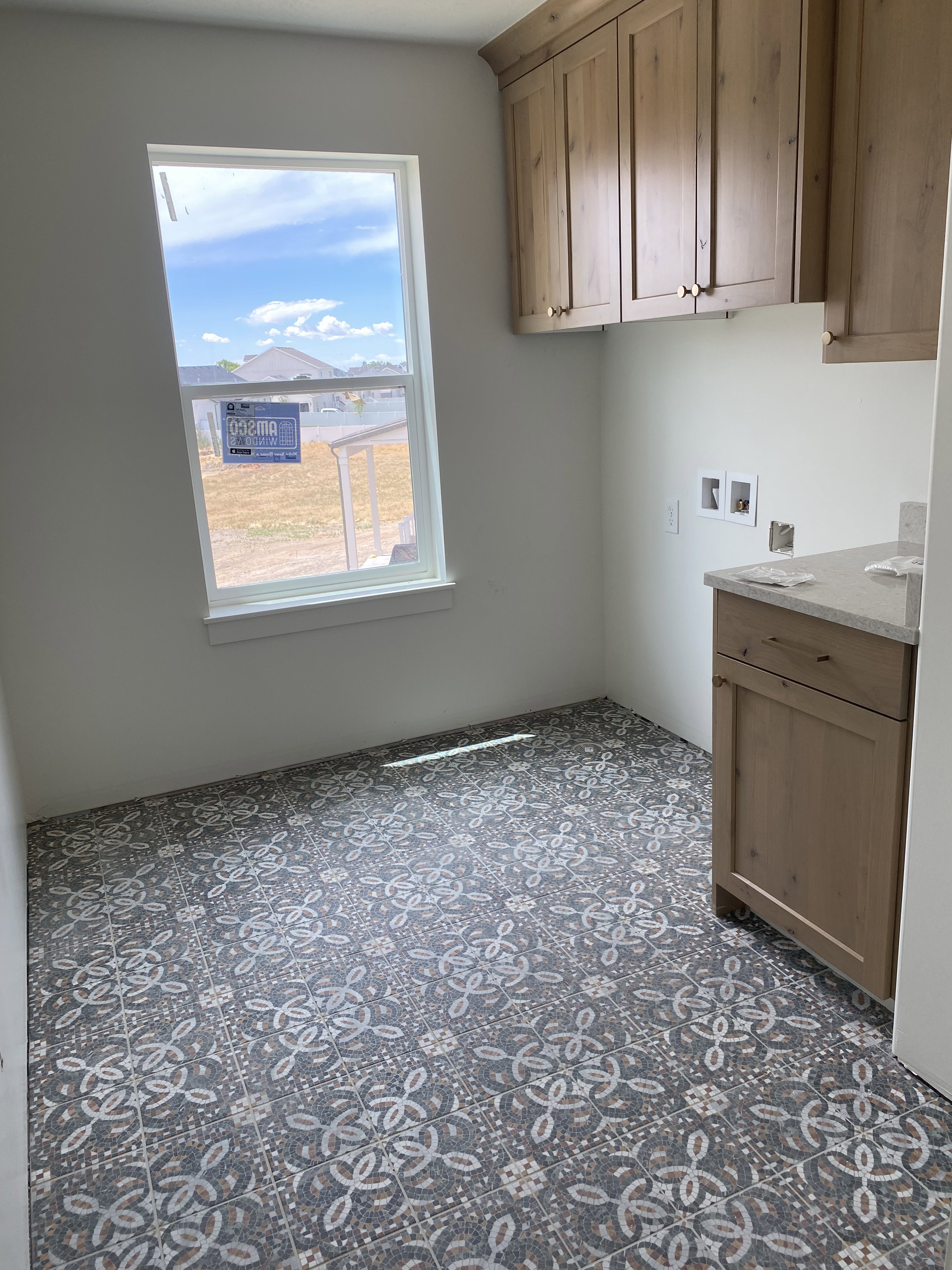 Laundry room featuring patterned tile flooring, light wood-finish cabinetry, a single window, and a light-toned countertop